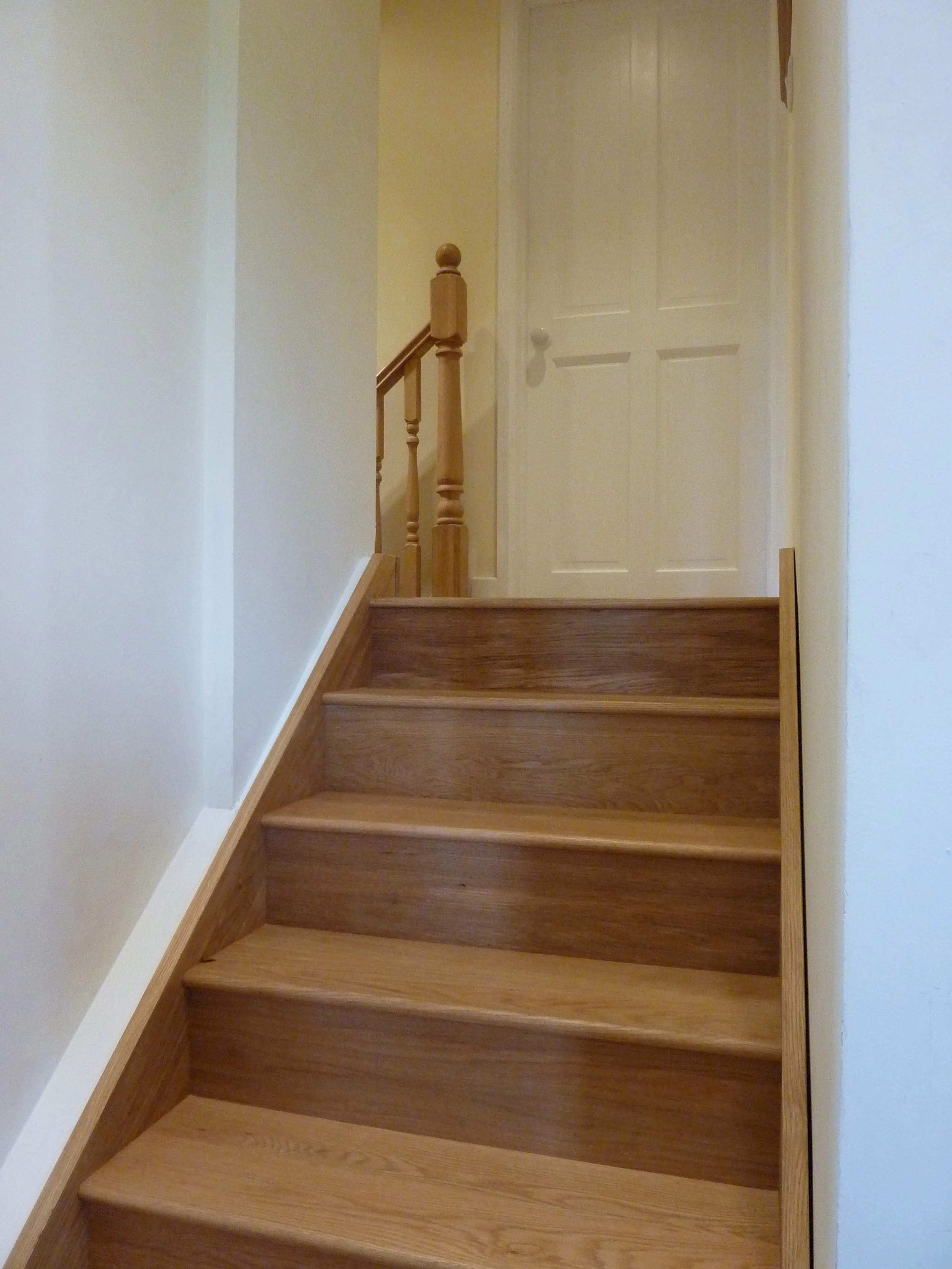 Wooden staircase with four steps leading to a closed door at the top, with a wooden banister on the left side.