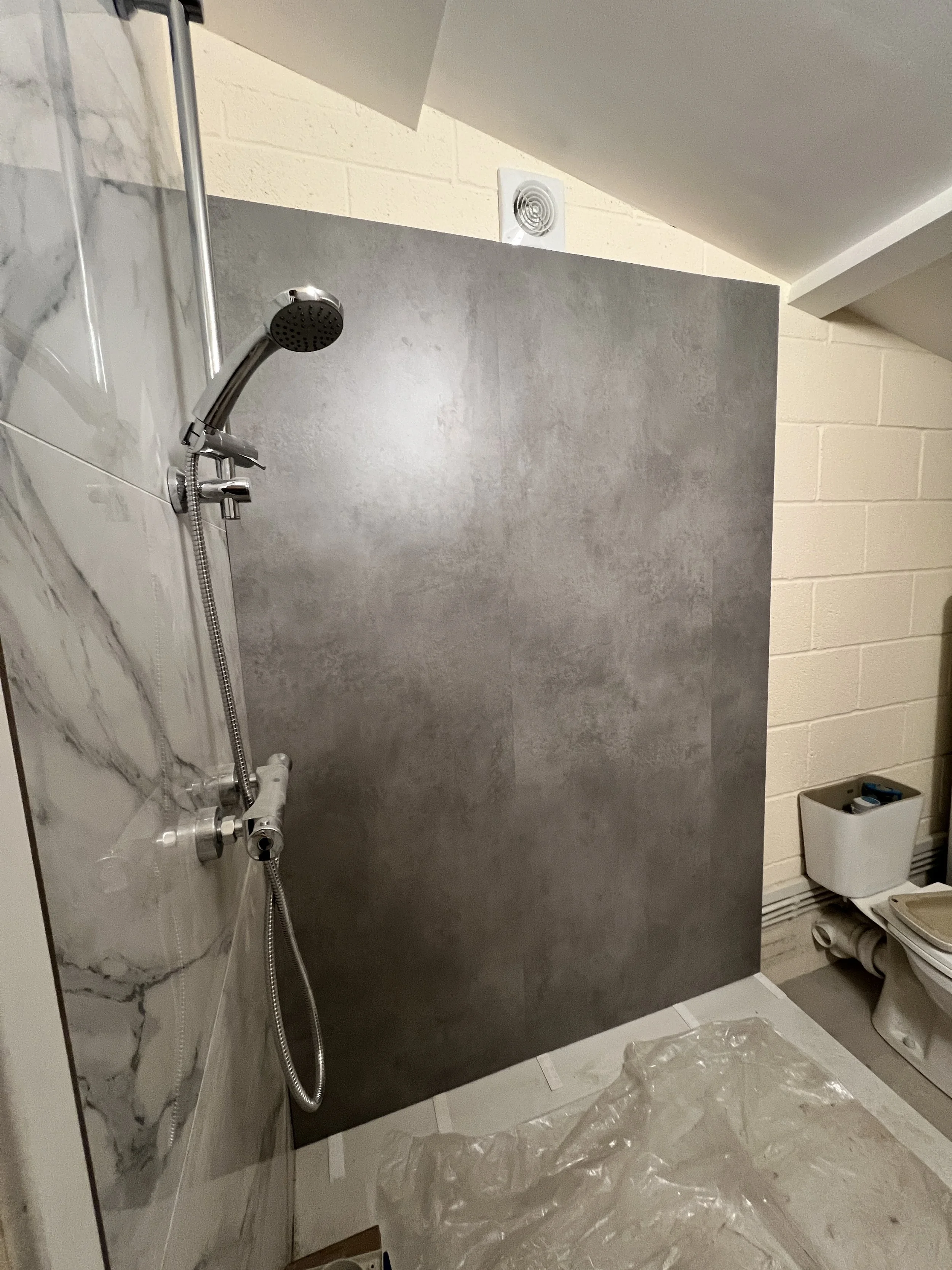 Interior of a bathroom with a shower area that has a grey concrete panel, a wall with marble-like tiles, a handheld showerhead, and an exposed toilet with plumbing and a trash bin nearby.