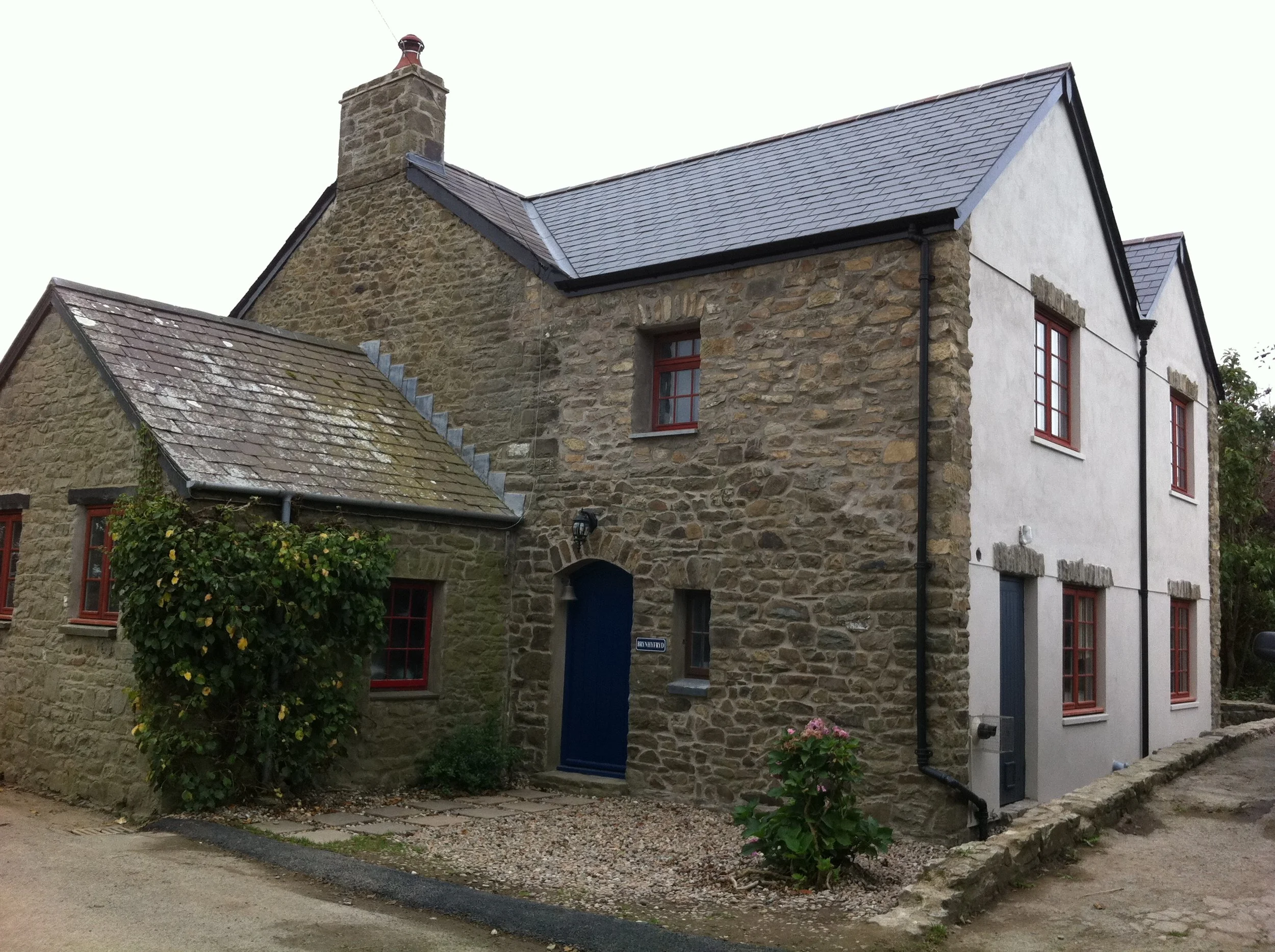 A stone house with a slate roof and red-trimmed windows, a chimney, and a blue door, with a garden and gravel pathway in the front.
