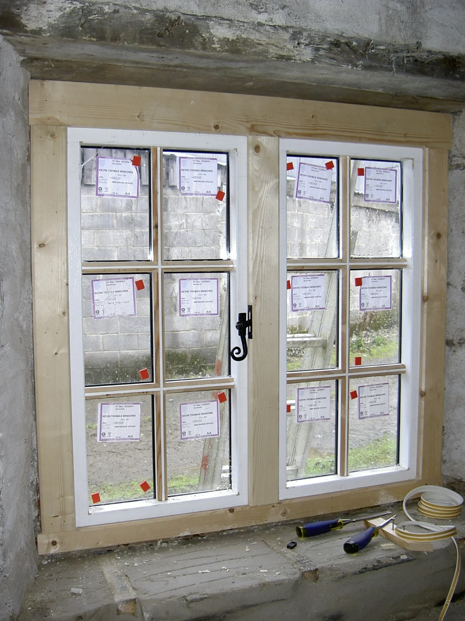A newly installed white window with multiple small glass panes in an unfinished room. The window is surrounded by a wooden frame, and construction tools and materials are on the floor.