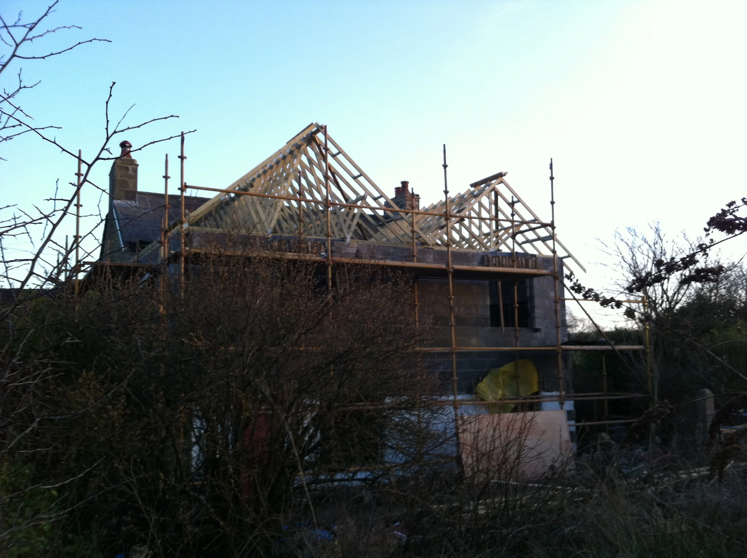 A house under construction with wooden roof trusses, surrounded by scaffolding and bare trees.