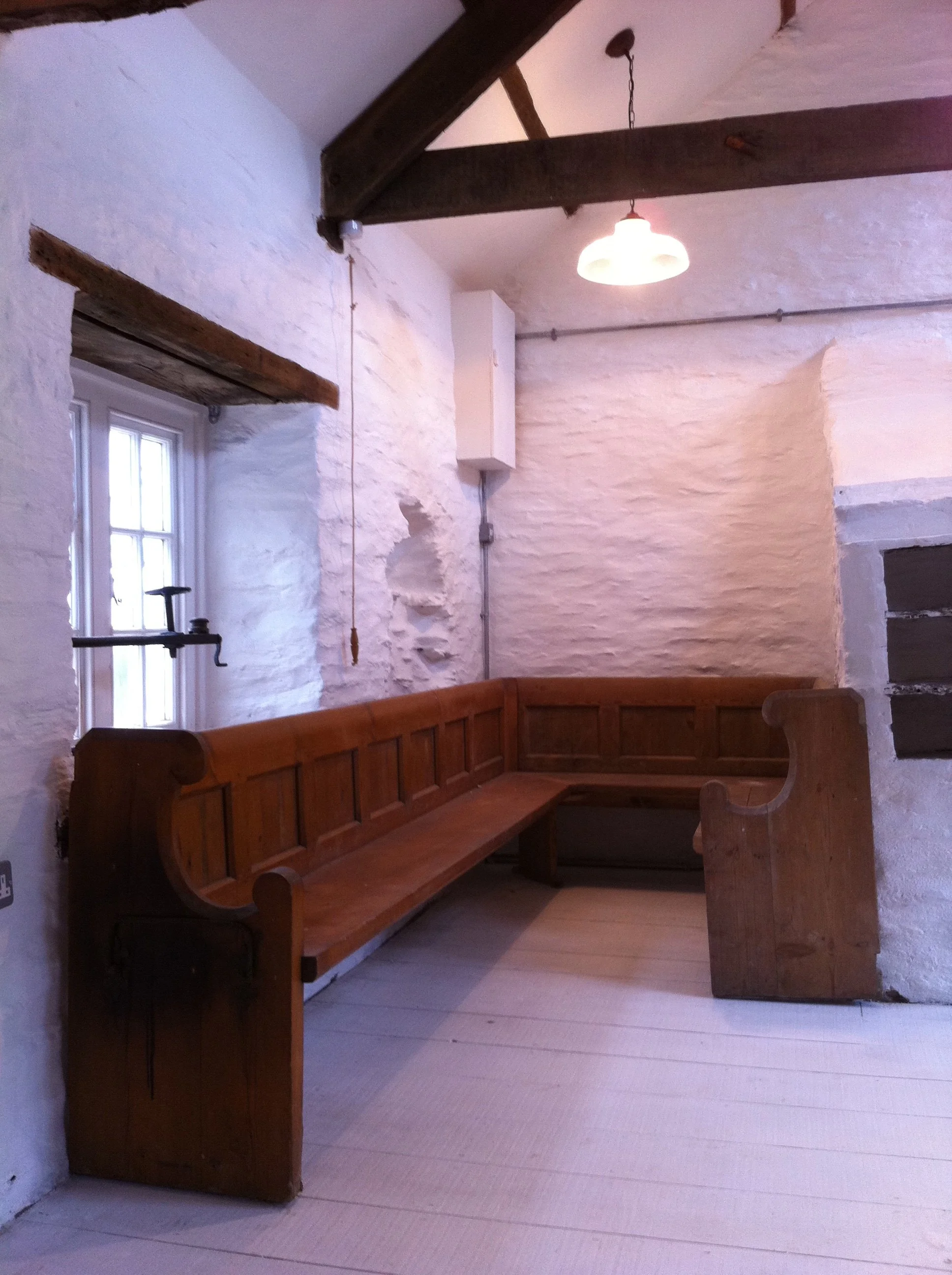 A corner of a room with lime wash painted stone walls, original A frames on the ceiling, a bespoke wooden bench with high sides, a small window, and a hanging light fixture in an airbnb