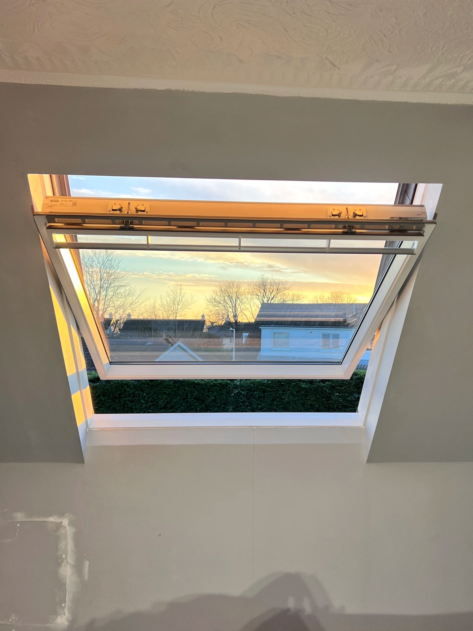 An open attic skylight window showing a sunset sky with trees and rooftops below.
