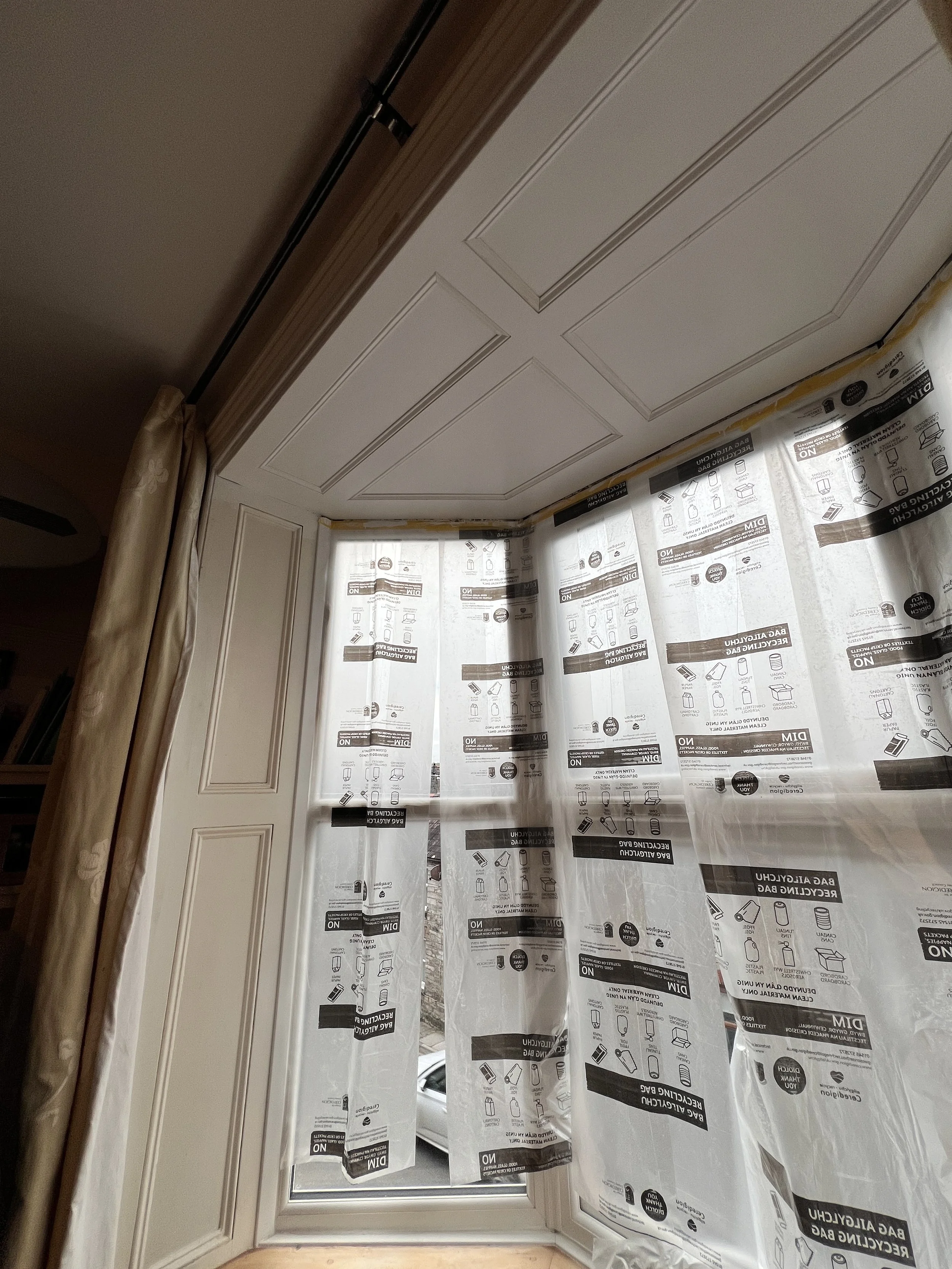 Room with window covered by plastic and tape, indicating painting or renovation work, with crown molding and drapery.