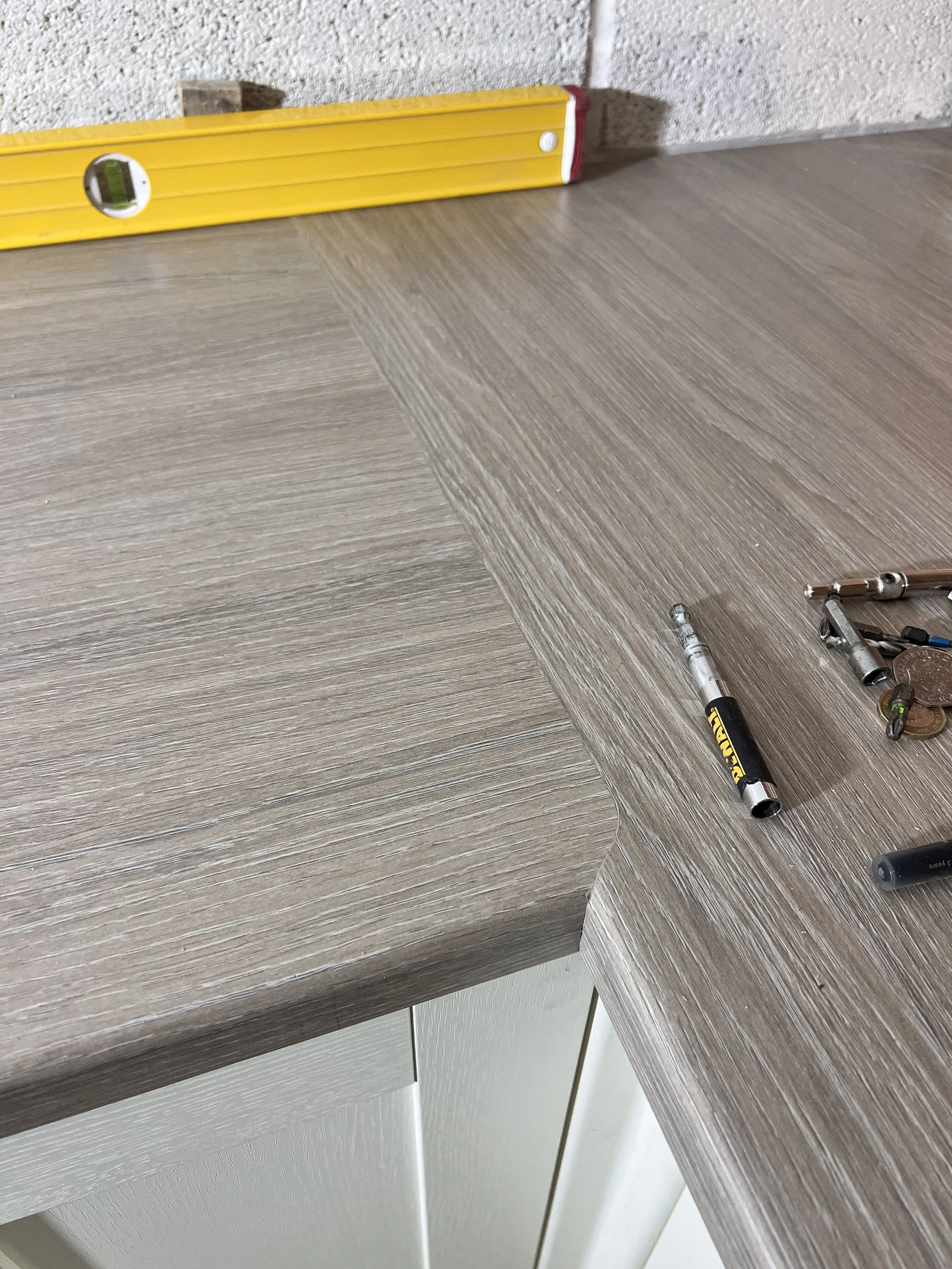 Close-up of a wooden countertop with a carpenter's level and some small tools lying on it. The corner of the countertop is rounded, and a wall with a textured surface is visible.