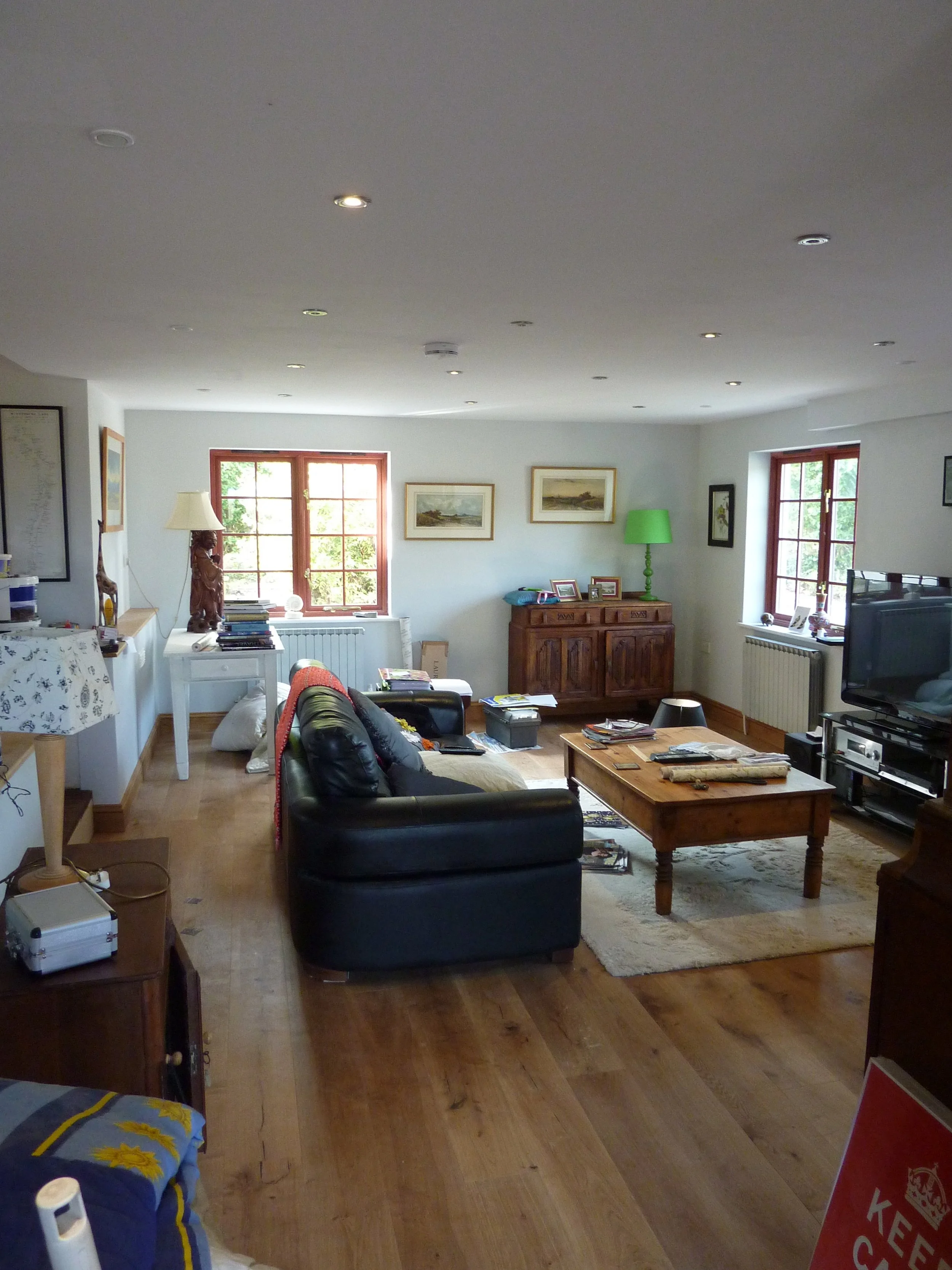 Living room with two windows, artwork on the wall, a wooden cabinet with picture frames and a green lamp, a black couch, and a wooden coffee table, with a television on the right, and various scattered items.