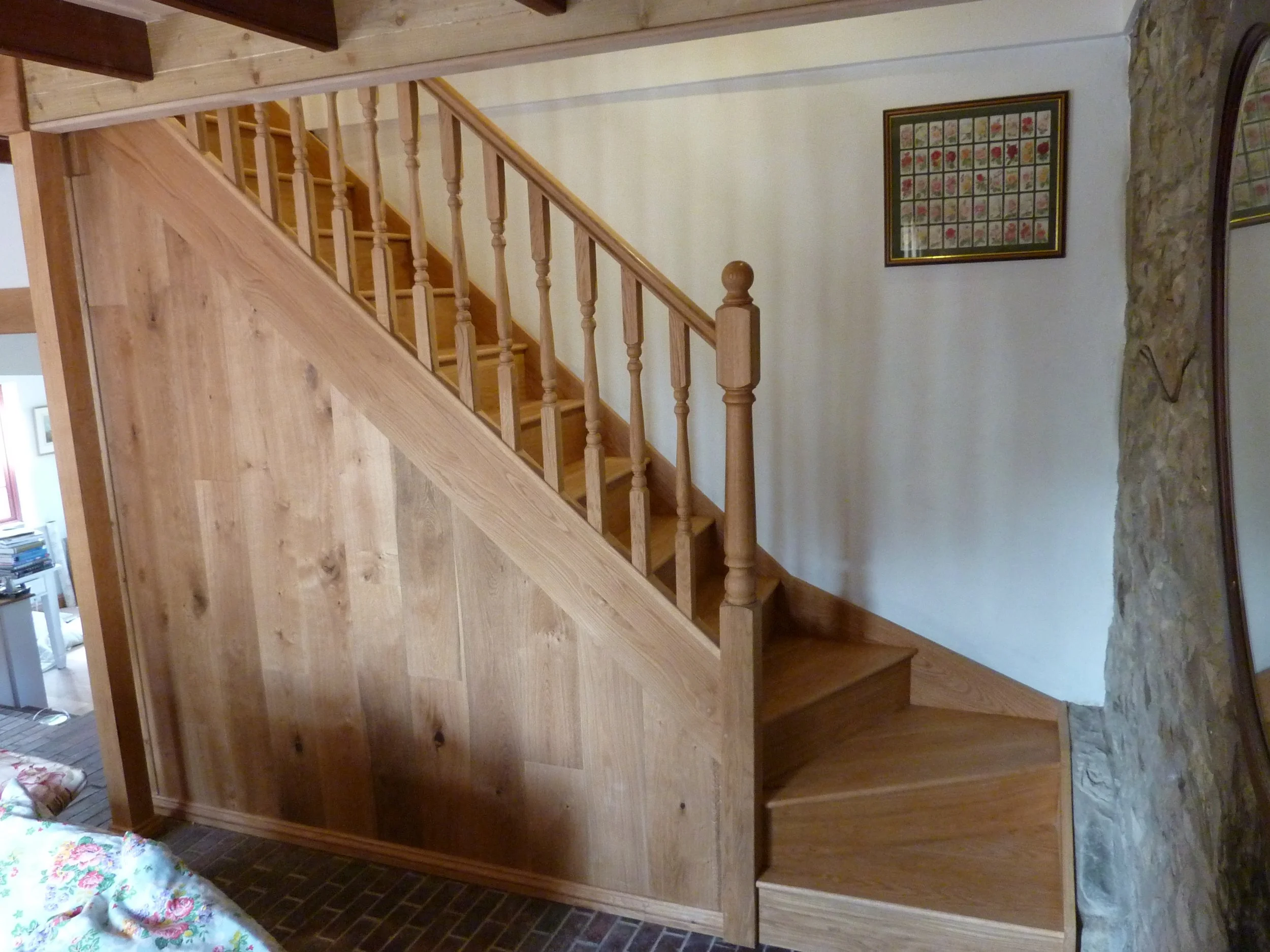 A wooden staircase with a railing inside a house, with framed artwork hanging on the wall adjacent to the stairs, and a stone accent wall near the landing.