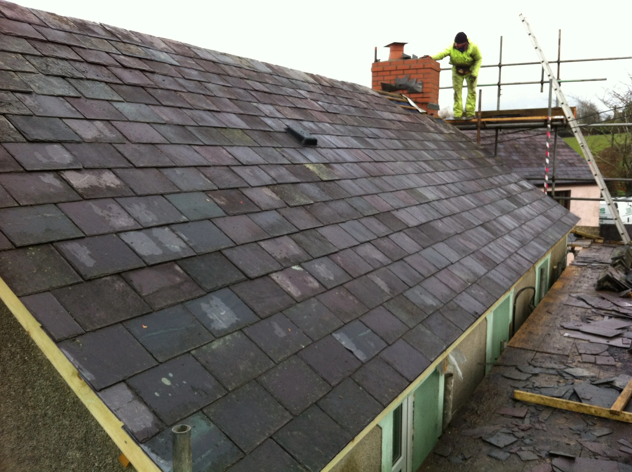 A worker in bright yellow safety overalls repairing a slate roof with scaffolding and a ladder in the background.