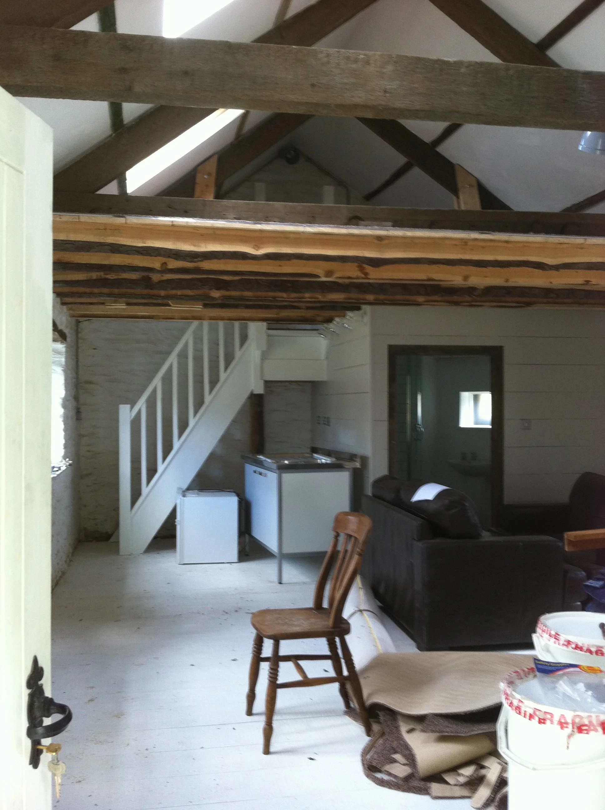Interior of a house under renovation with exposed wooden ceiling beams, unfinished floors, a staircase with a white railing, some furniture including a black couch, a wooden chair, and various renovation supplies.