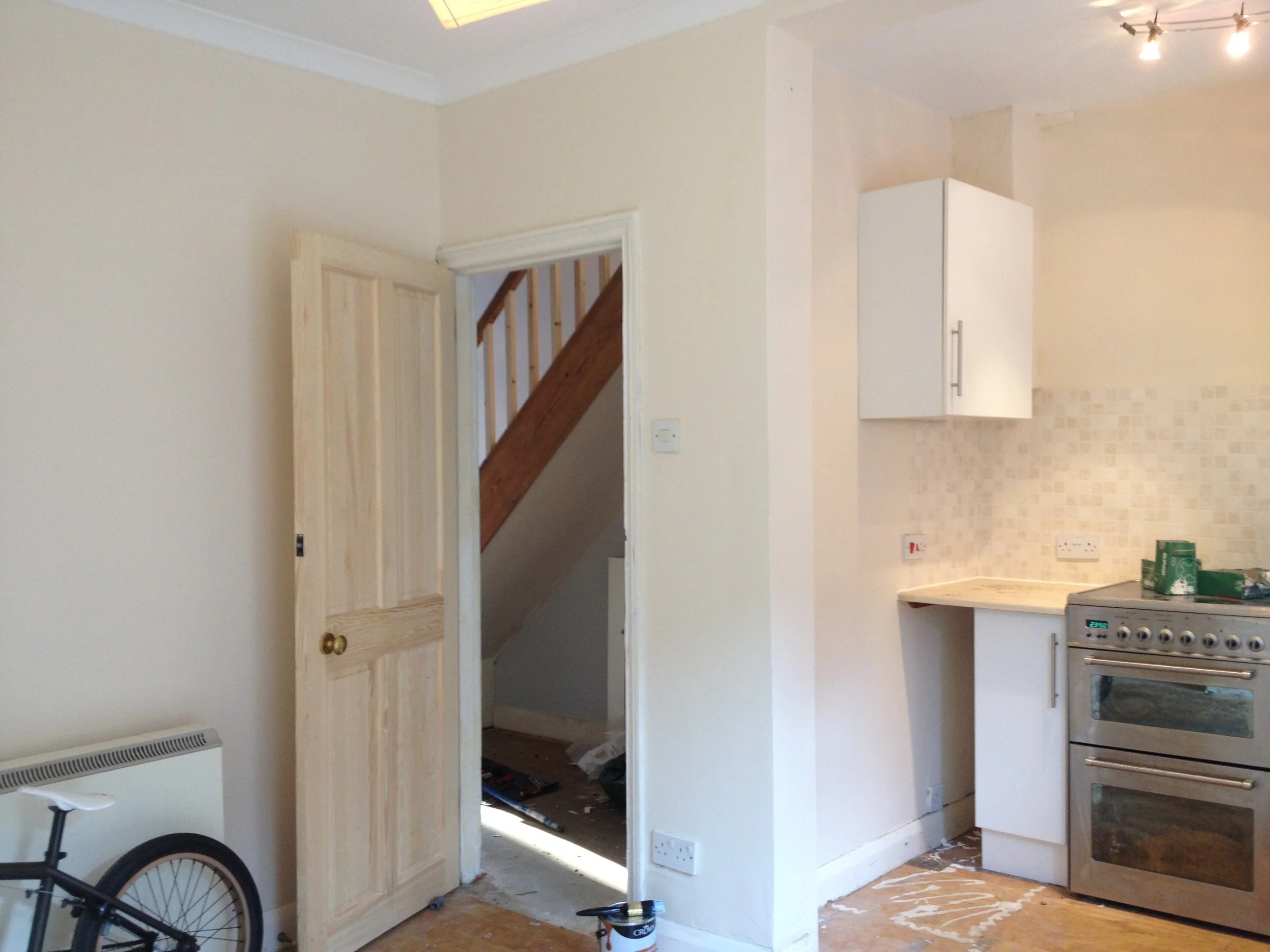 Interior of a kitchen with white walls, wooden cabinetry, and an open doorway leading to a staircase. There is a stove on the right, and a small corner of a black bicycle is visible in the foreground. The floor is covered with plastic and some tools.