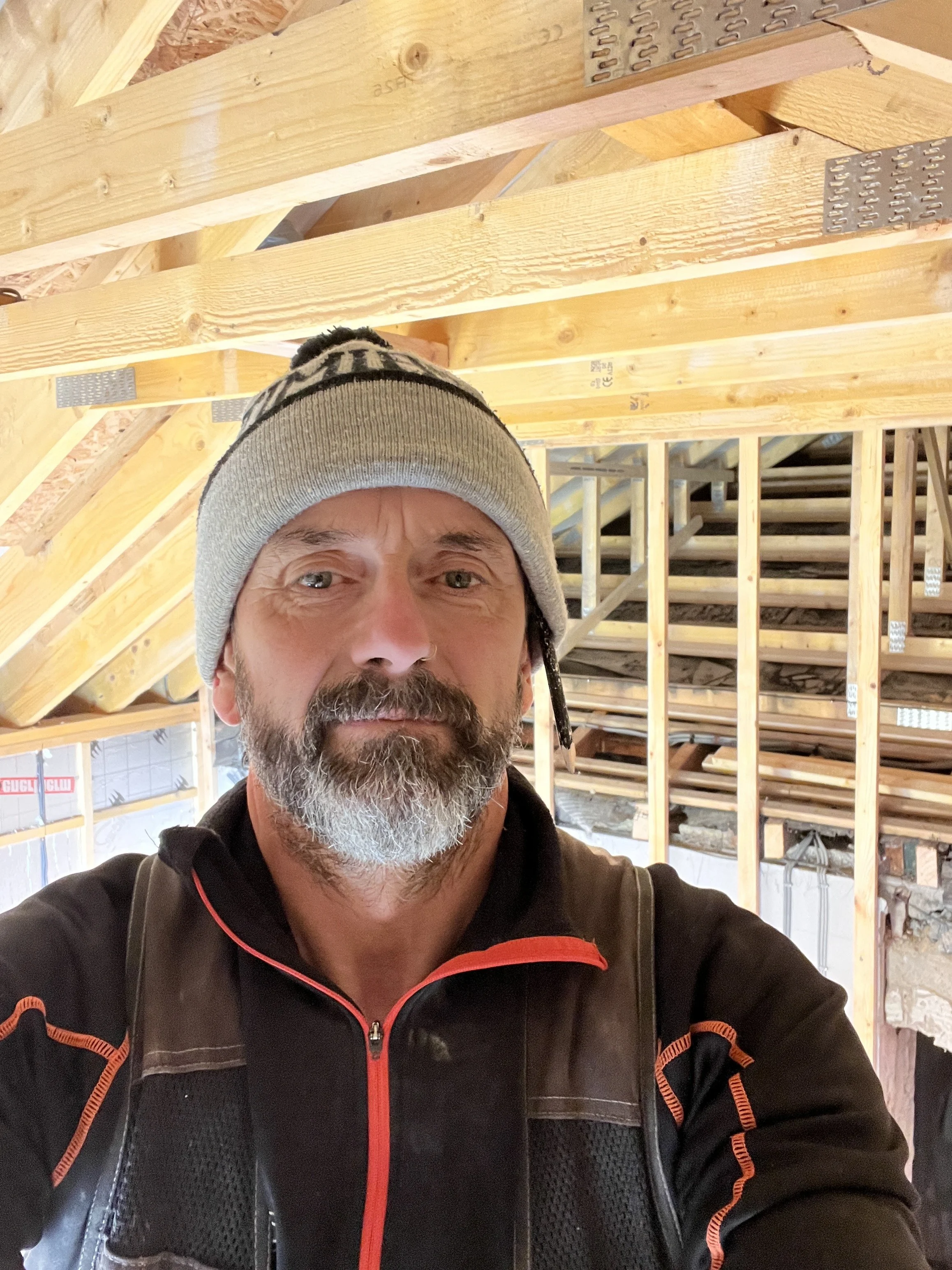 A man with a gray beard and mustache wearing a gray beanie hat and a black zip-up jacket with orange accents, taking a selfie at a construction site with exposed wooden framing in the background.