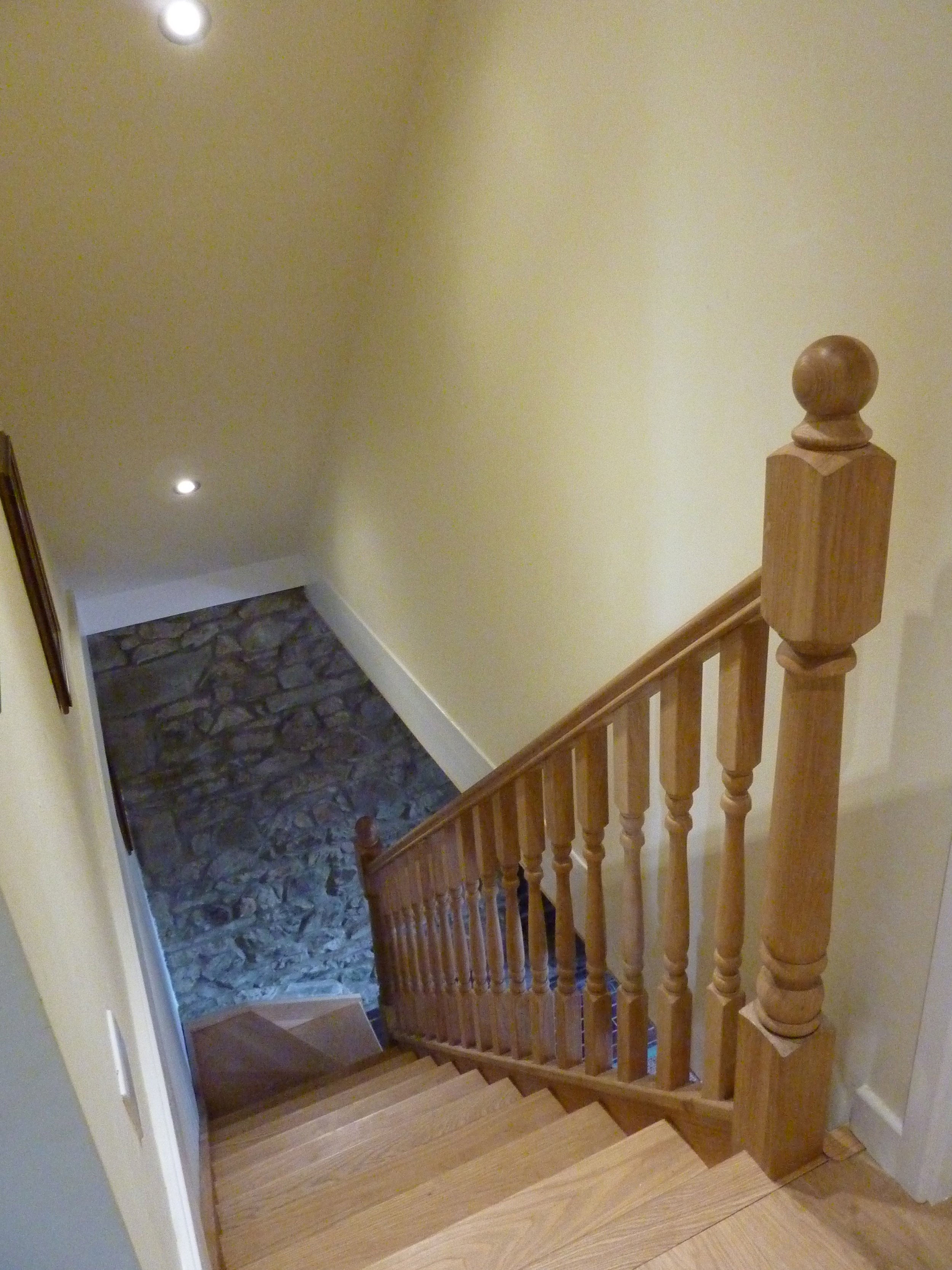 A wooden staircase viewed from the top, leading down to a stone floor and a yellow wall on the right side.