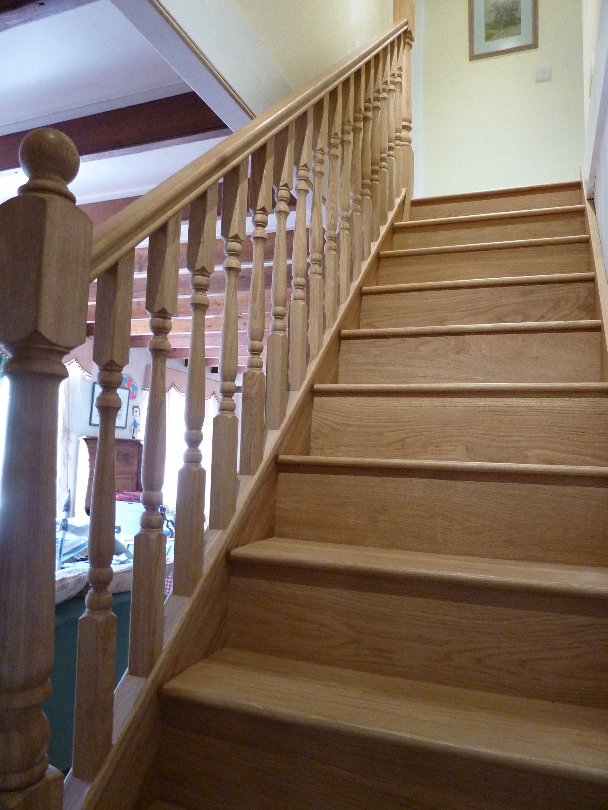 Wooden staircase with balustrade inside a house, leading upstairs to a hallway with paintings and a door.