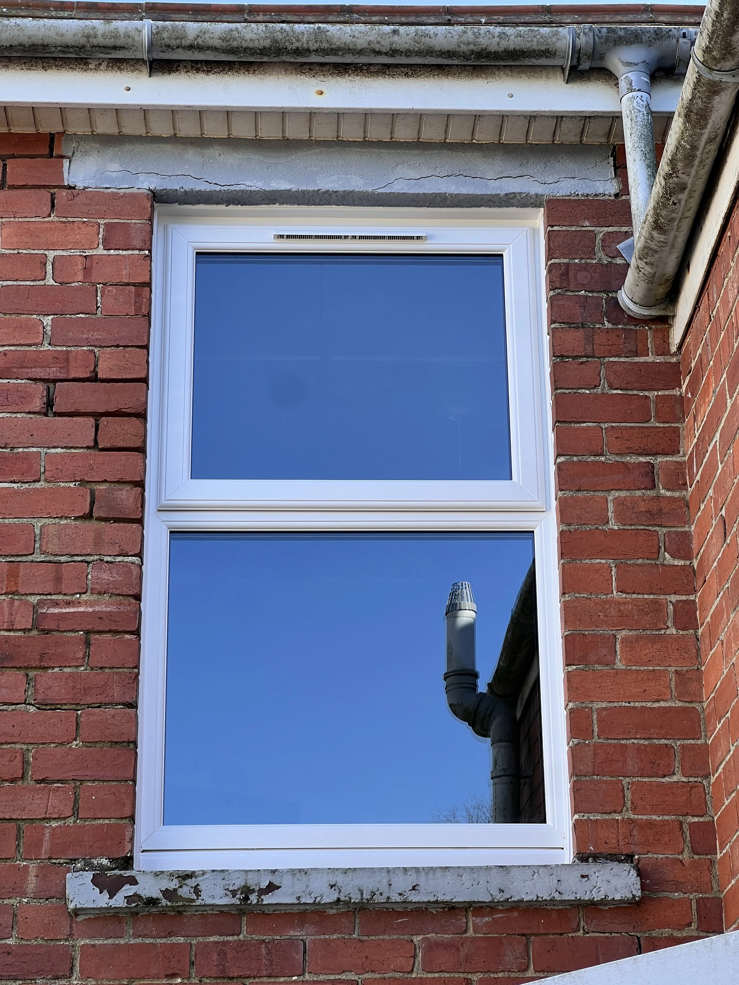 Close-up view of a brick building with a white-framed window and visible cracks in the wall above the window. There are drainage pipes and a chimney in the scene.