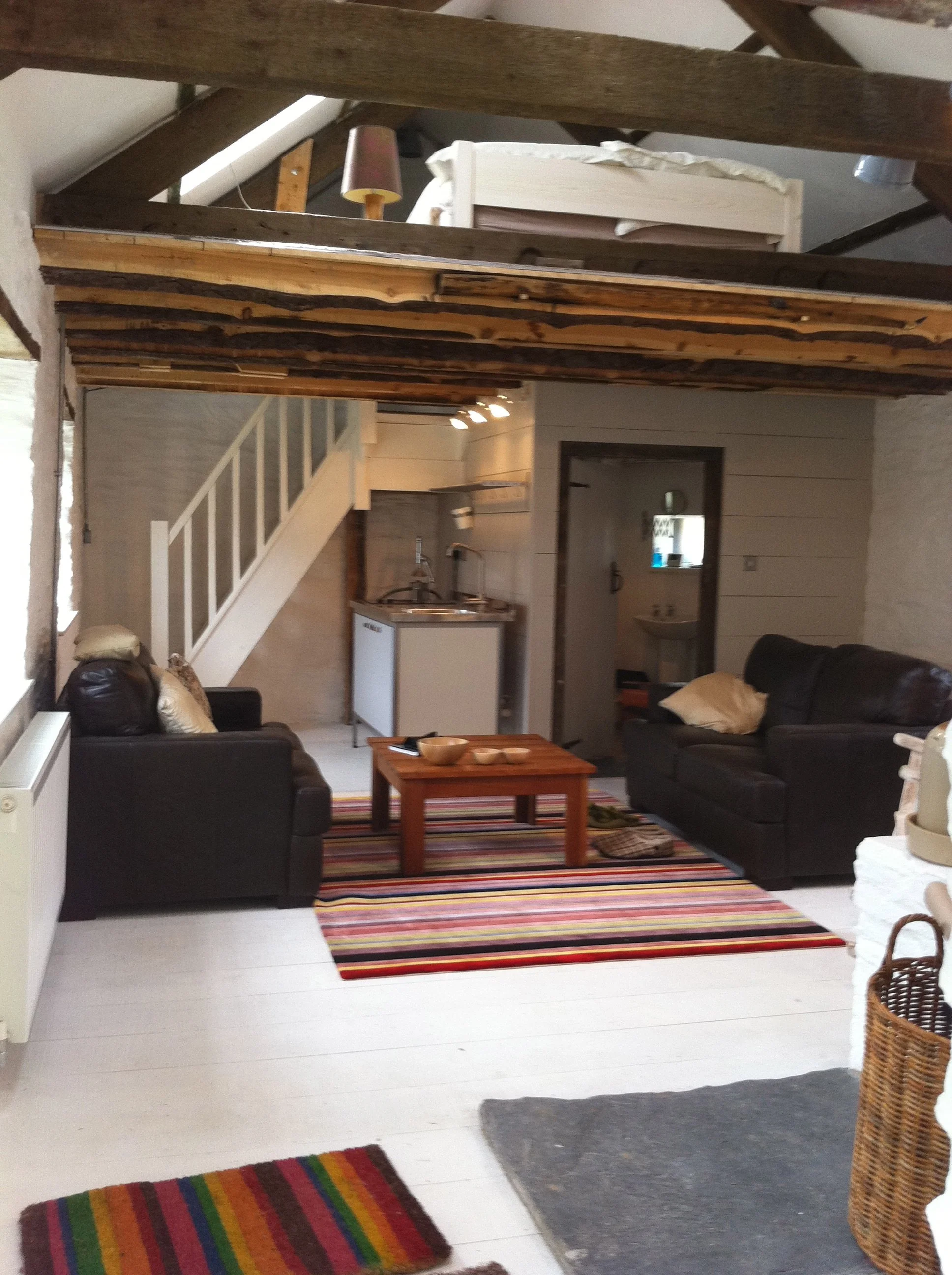 Living room with black sofas, a wooden coffee table, and colorful striped rugs, with a staircase and kitchen area in the background, and a loft with wooden beams overhead.