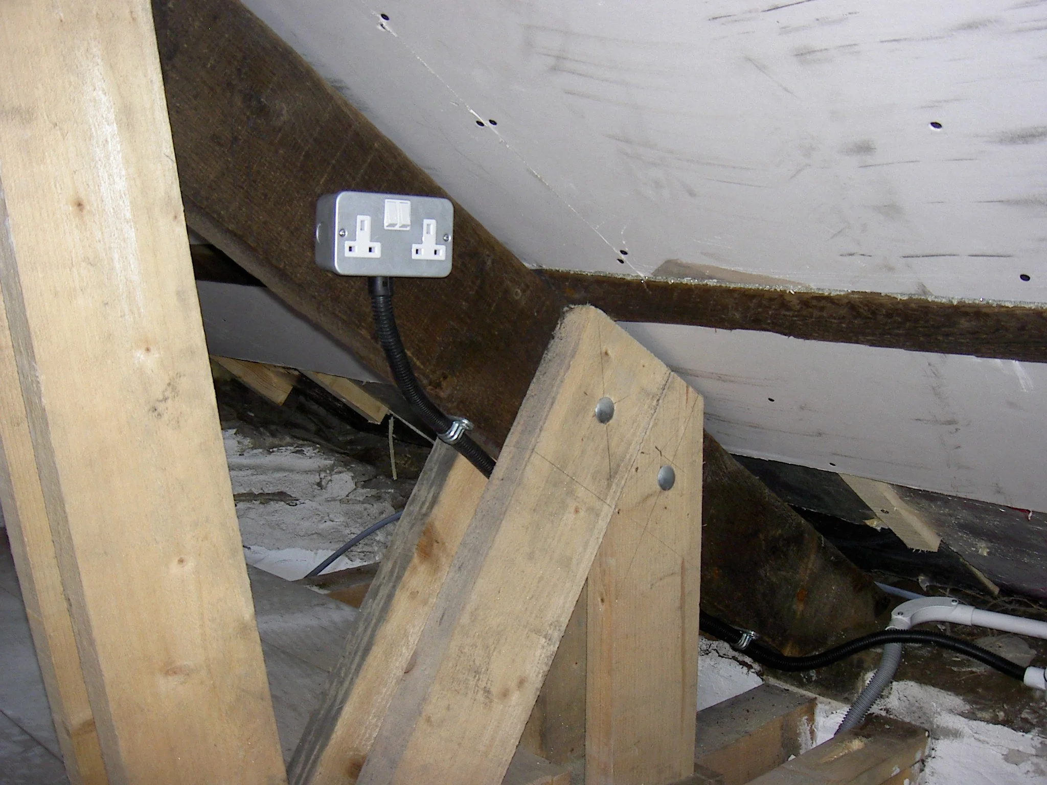 An unfinished attic corner with wooden framing and electrical outlets, insulation, and wiring visible.