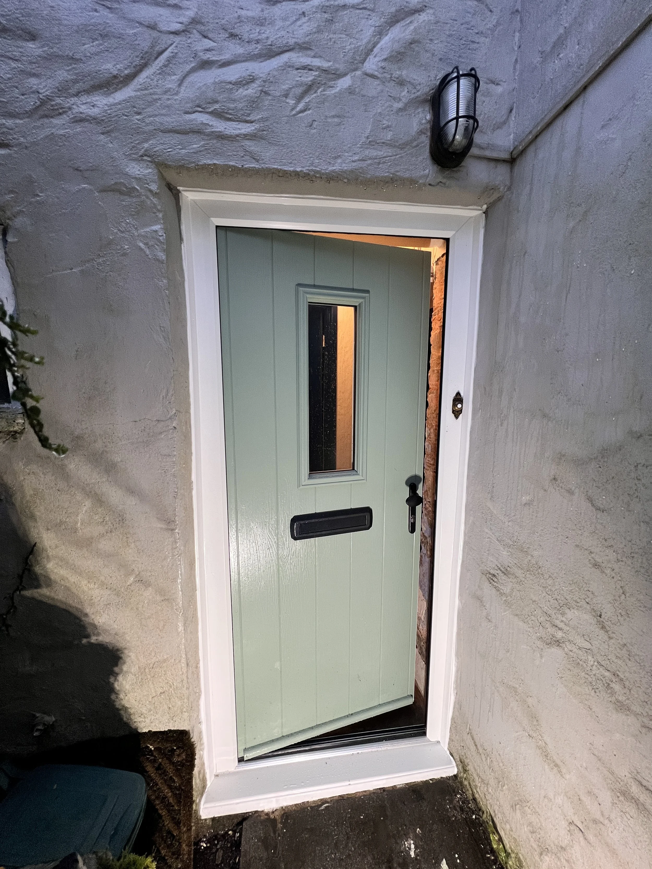 A green front door with a black handle and mail slot, set in a white trim frame. There is a window in the door, revealing a warmly lit interior. A black outdoor wall light is mounted above and to the right of the door. The surrounding wall is made of