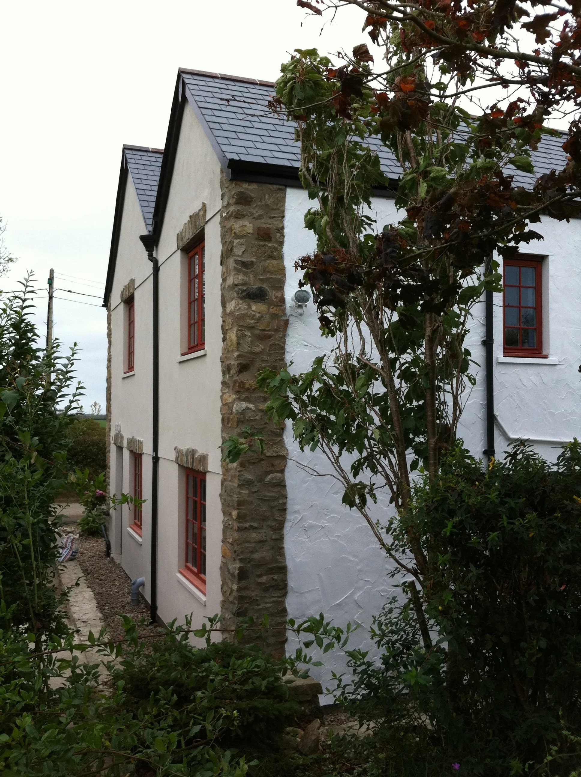 Side view of a house with white stucco walls, red window frames, a stone corner, and a slate roof, surrounded by greenery and trees.
