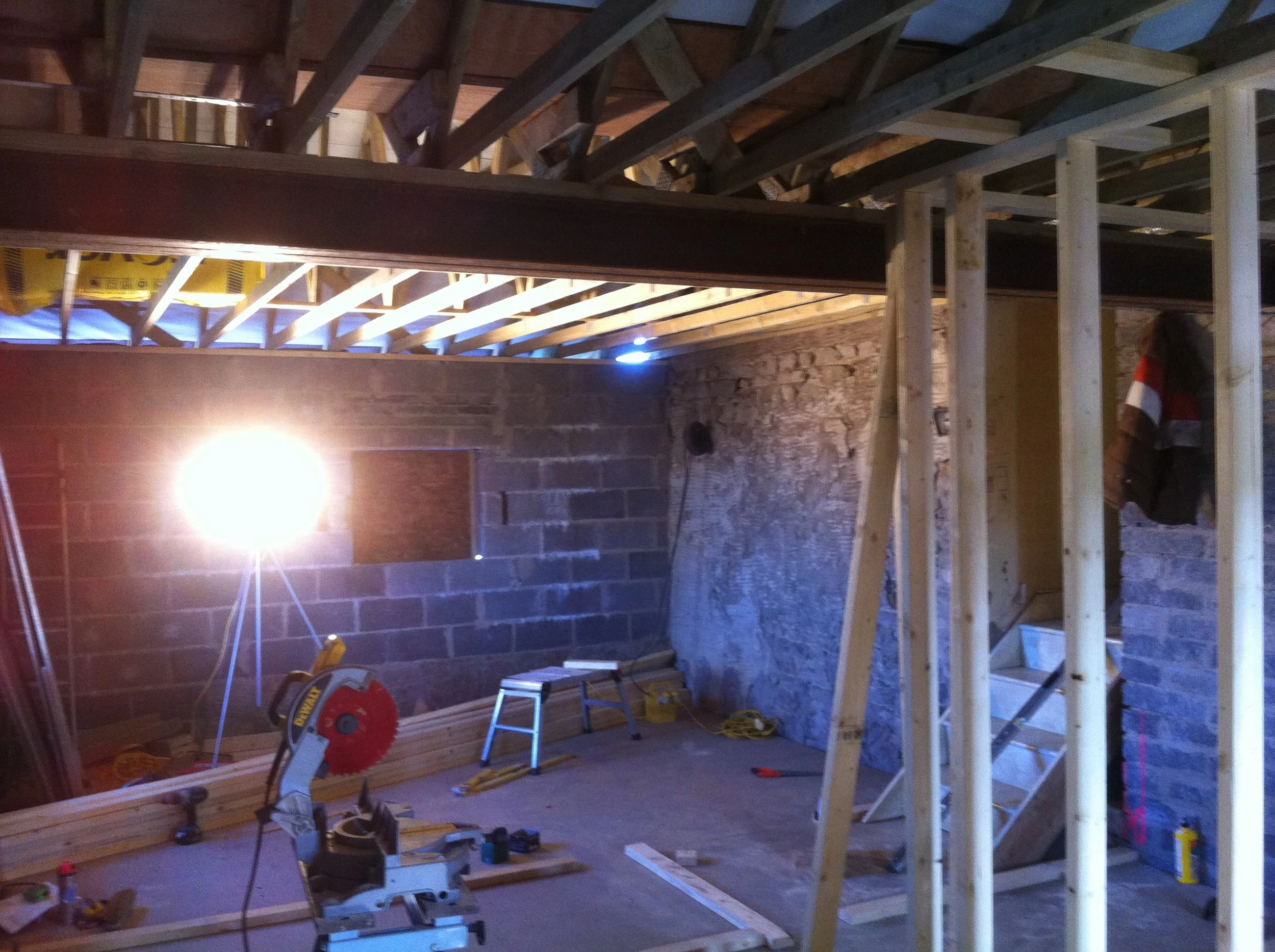 Interior of a building under construction with exposed roof trusses, a partially built interior wall with block work, stud walls and construction tools.