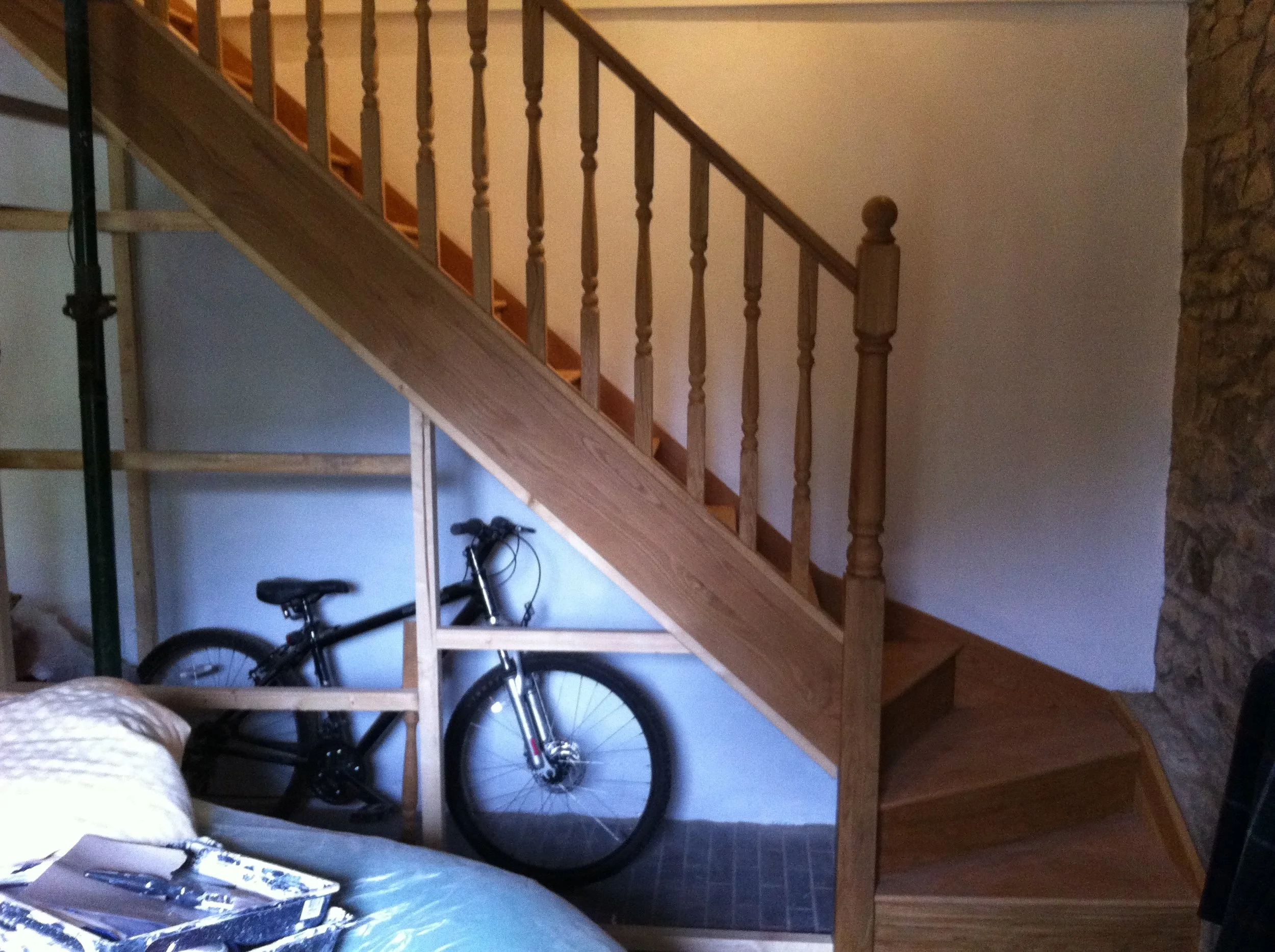 A wooden staircase with balusters leading to an upper level, adjacent to a stone wall, with a bicycle parked underneath and an art palette visible in the foreground.