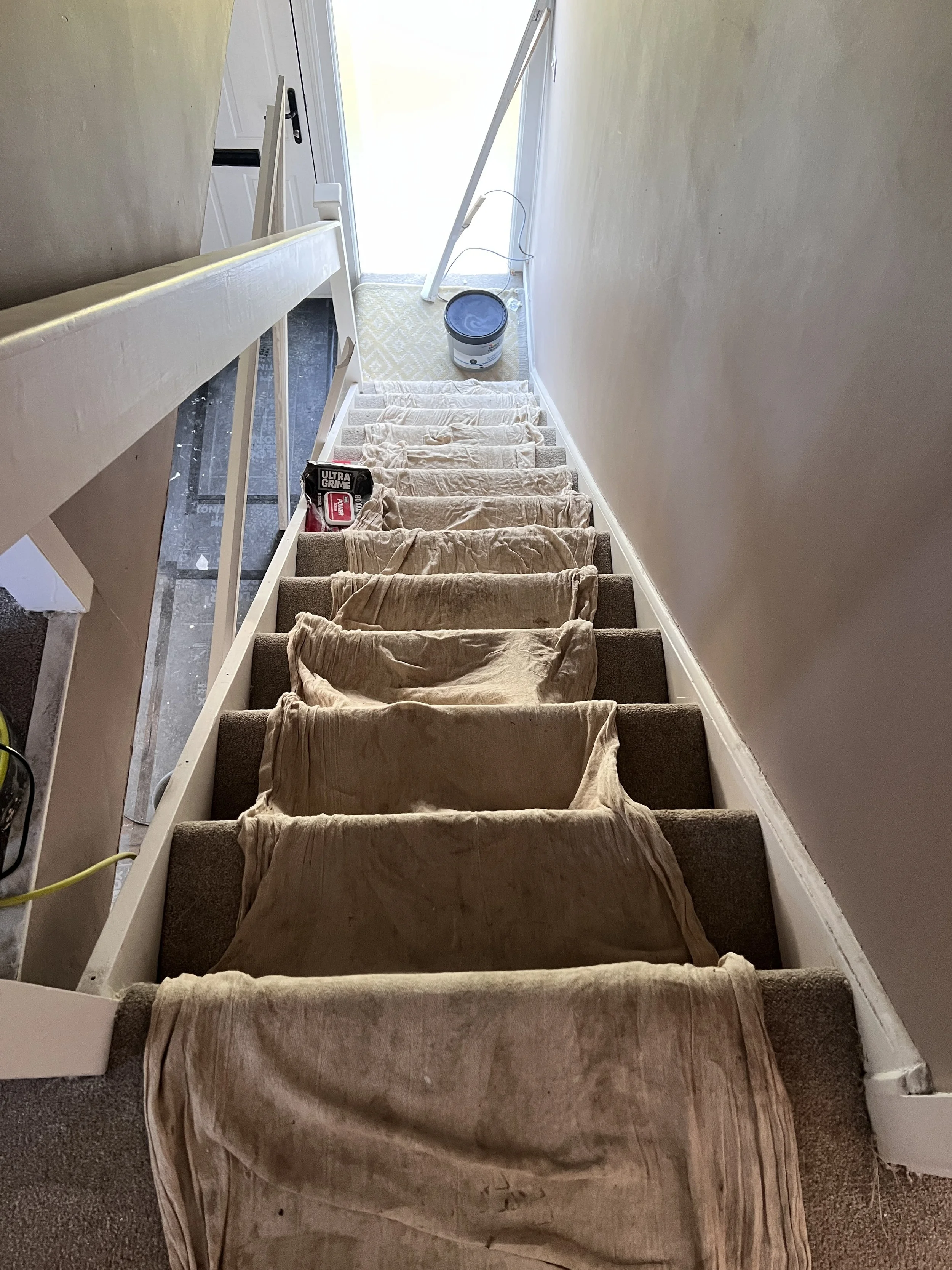 A staircase being painted with beige paint; paintcloth covers the steps, and painting supplies such as a paint roller tray, a paint roller, and a paint bucket are visible.
