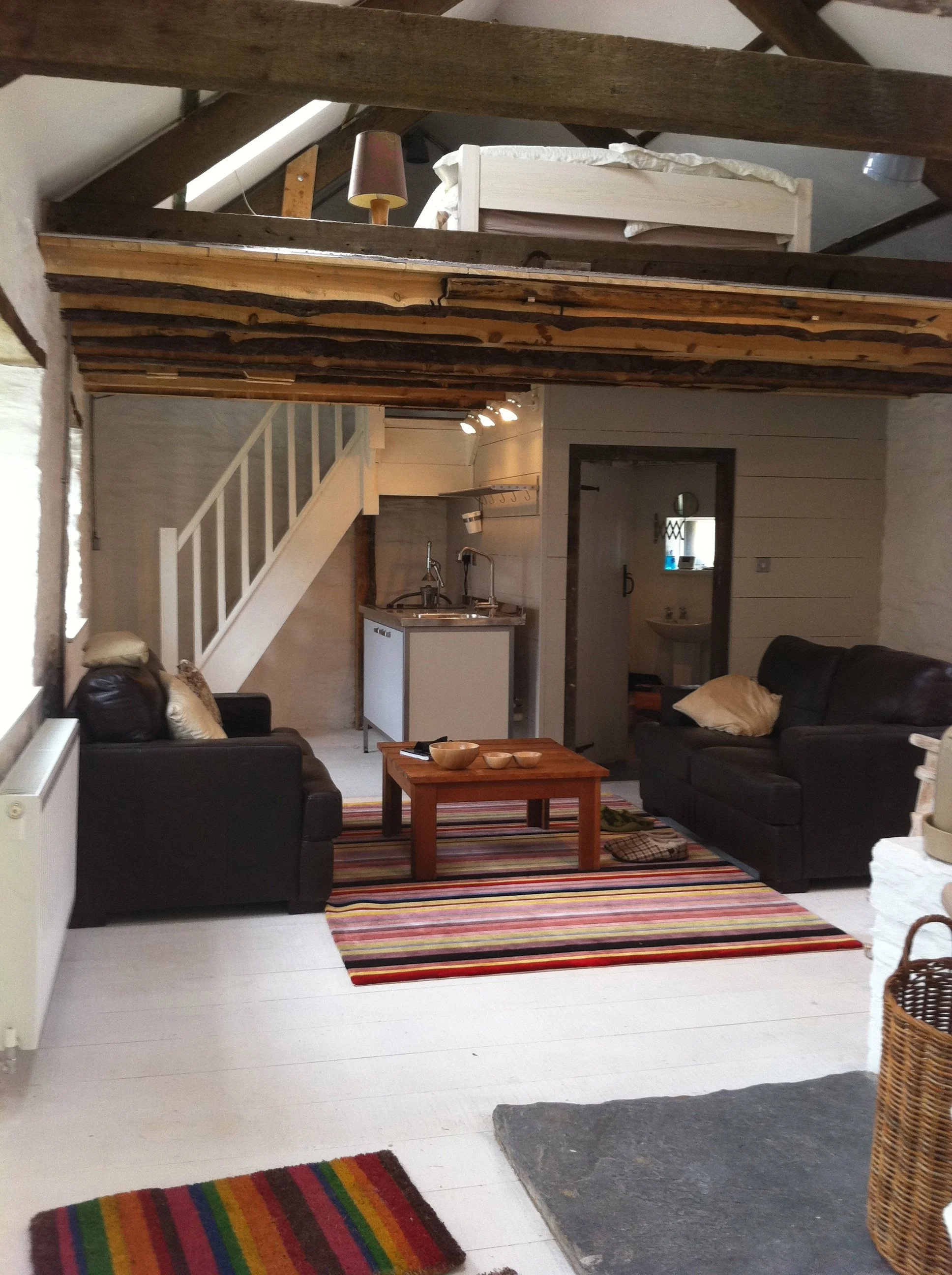 Living room with black sofas, a wooden coffee table, a colorful striped rug, white walls, a staircase, kitchen area with a small sink, and a lofted bed with a blanket and pillow.