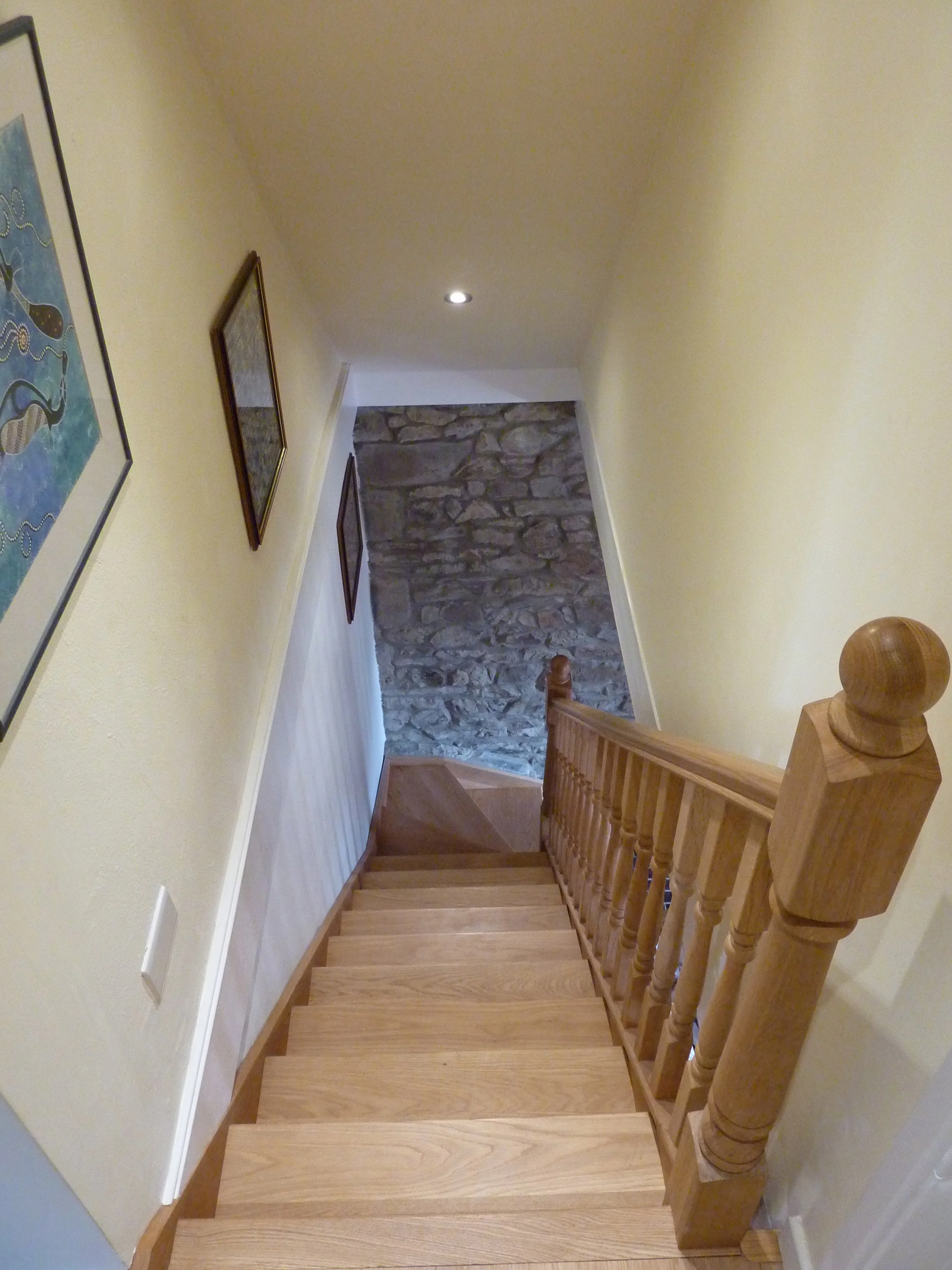 Wooden staircase leading down, with a stone wall at the bottom and framed artwork and a framed photo on the yellow wall.
