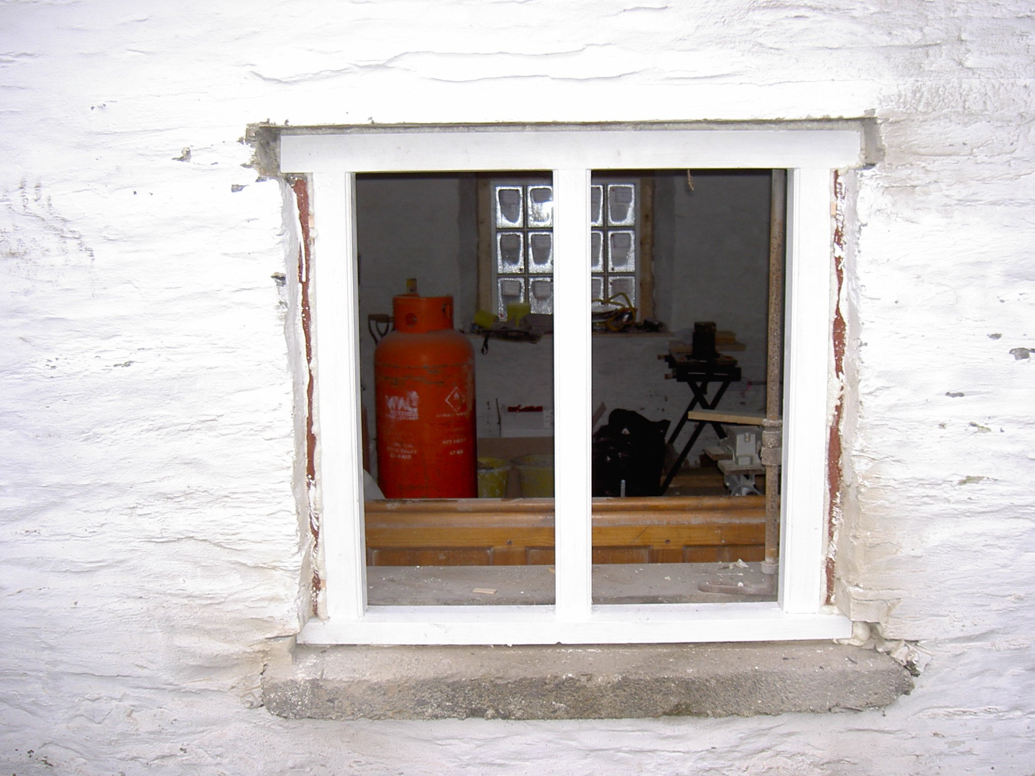 White brick wall with a rectangular window opening under construction, revealing a room inside with a large orange propane tank and construction tools.