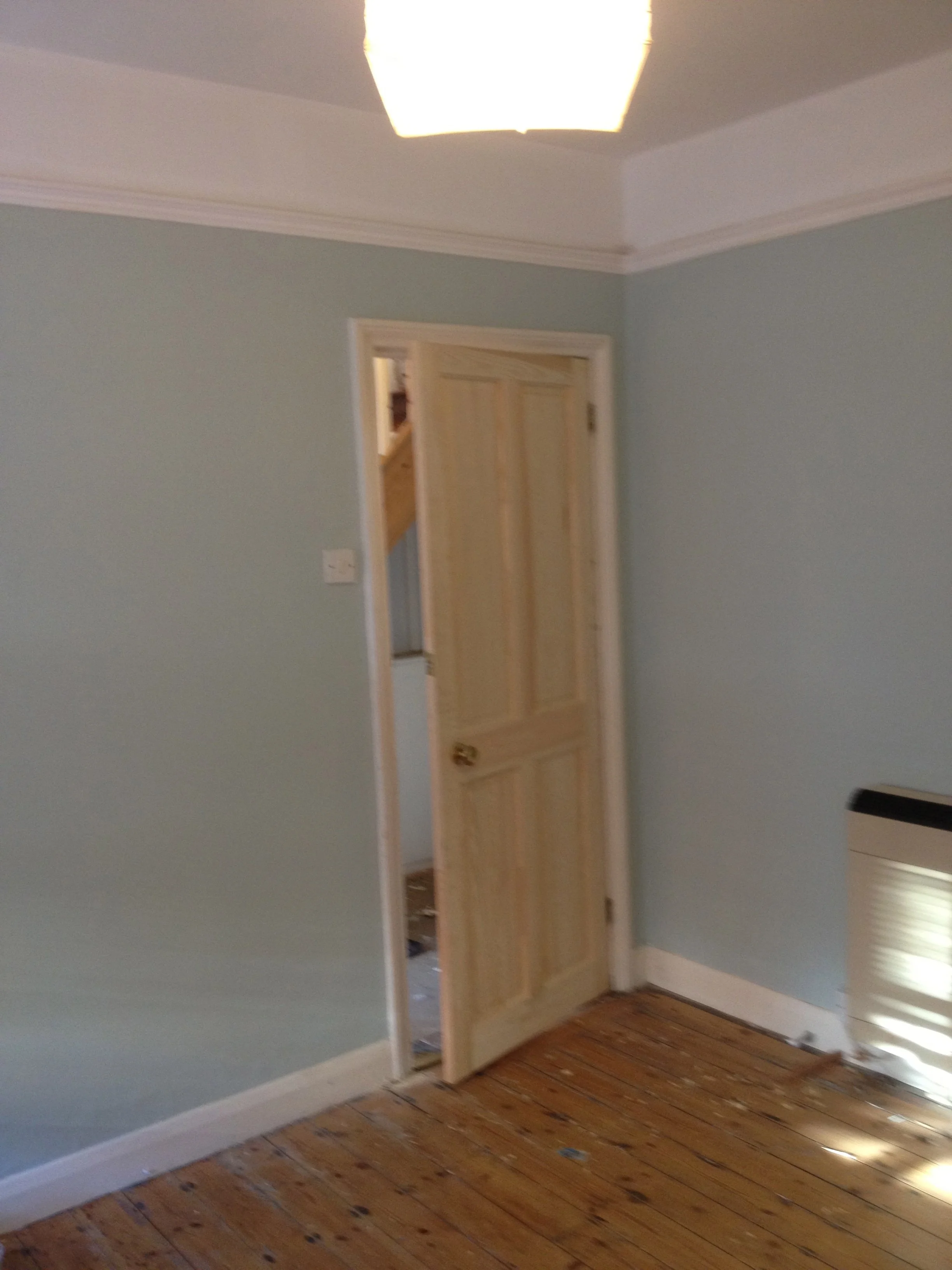 Interior of a room with light green walls, a wooden banister visible through an open door, a ceiling light, and wooden flooring.