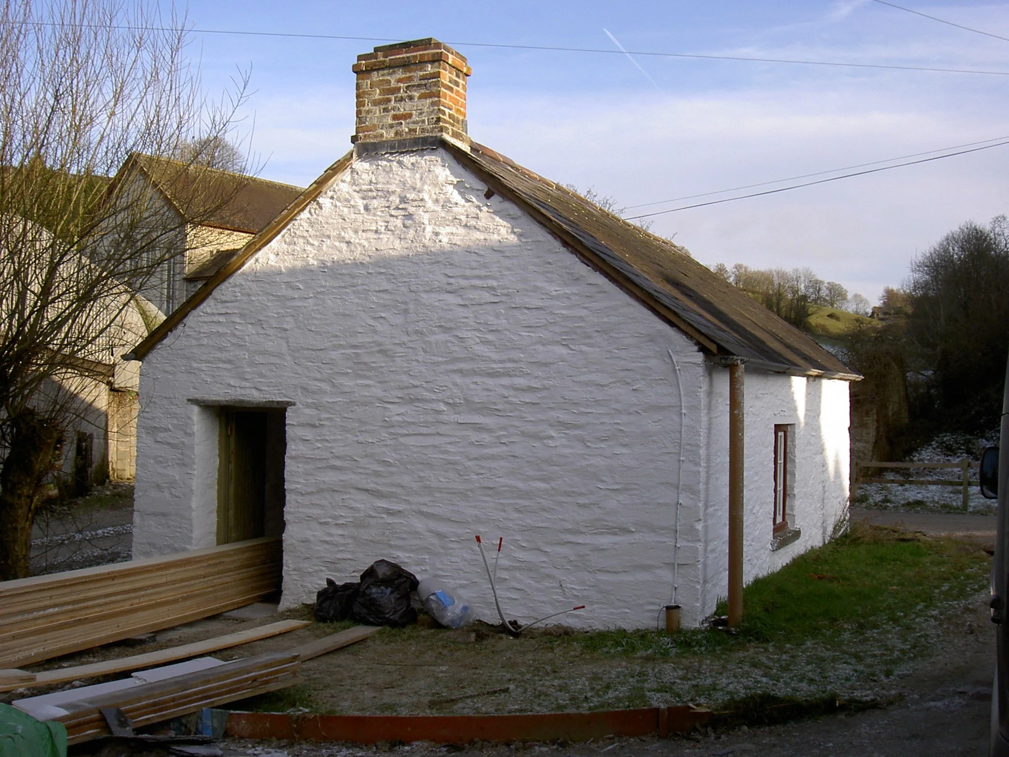 A white house under construction with a brick chimney, construction materials, and tools in the yard, trees in the background, and a blue sky with some clouds.