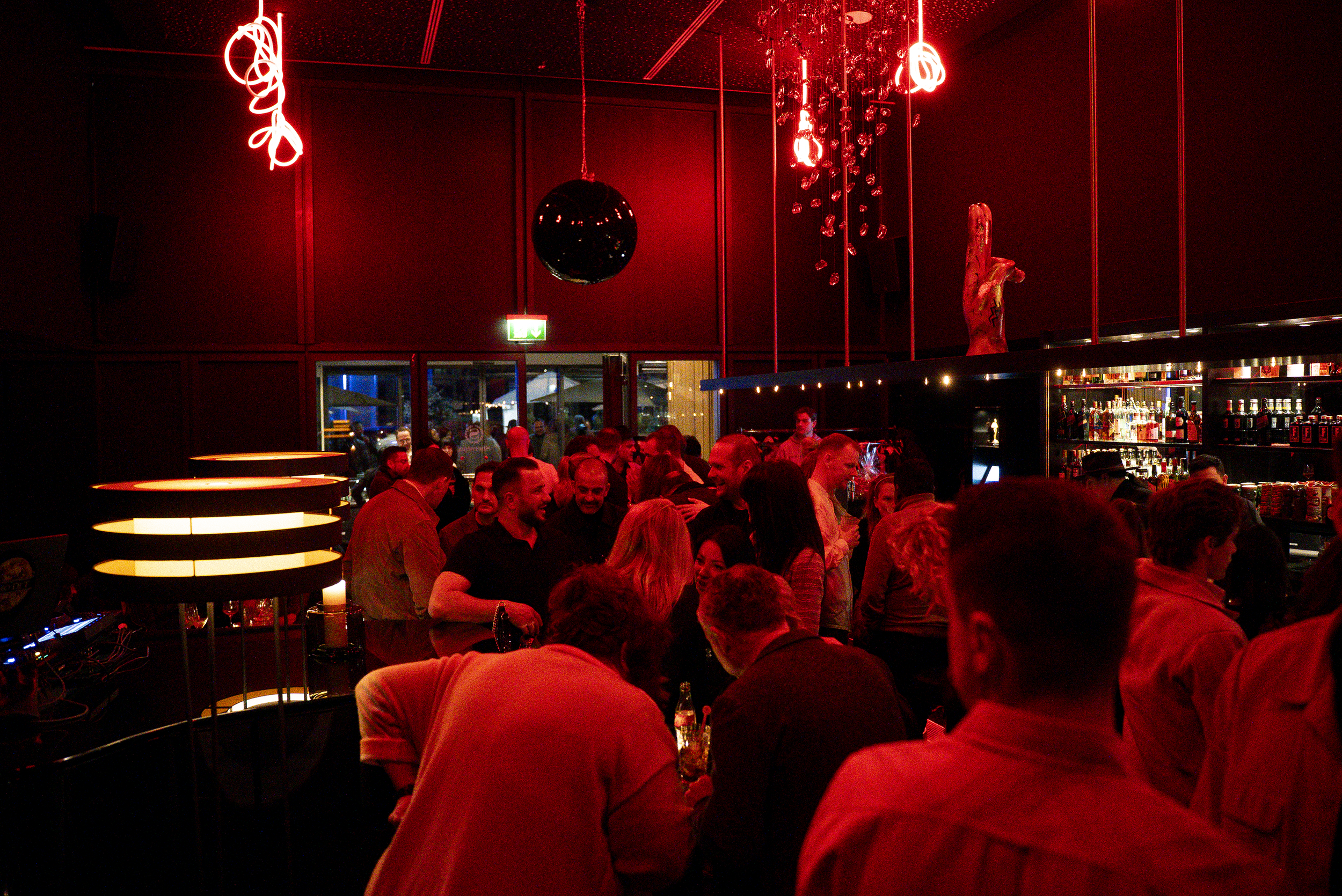 A crowded bar or nightclub with red lighting, people socializing, and bottles of alcohol on display behind the bar.