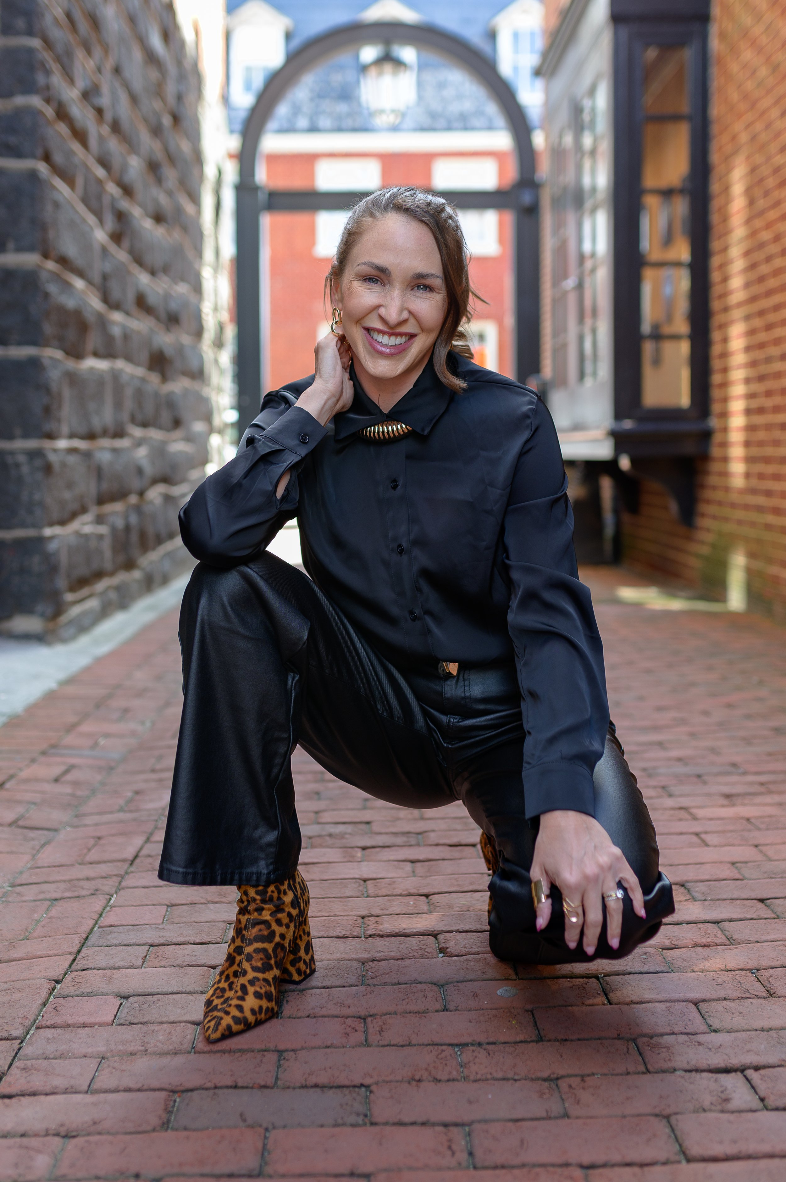 A woman in black leather clothing, leopard print boots, and leopard print pants crouching on a brick sidewalk. She is smiling, with her hand on her neck, and holding a black bag. The background shows buildings and an archway.