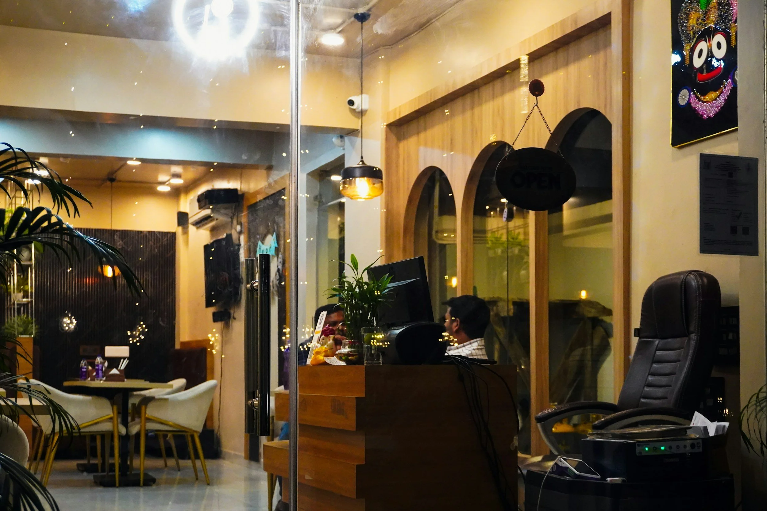 Interior of a cozy restaurant or cafe seen through a large glass window with a reception desk in the foreground, two people sitting and talking inside, decorated with plants and warm lighting, massage vending chairs in foreground.