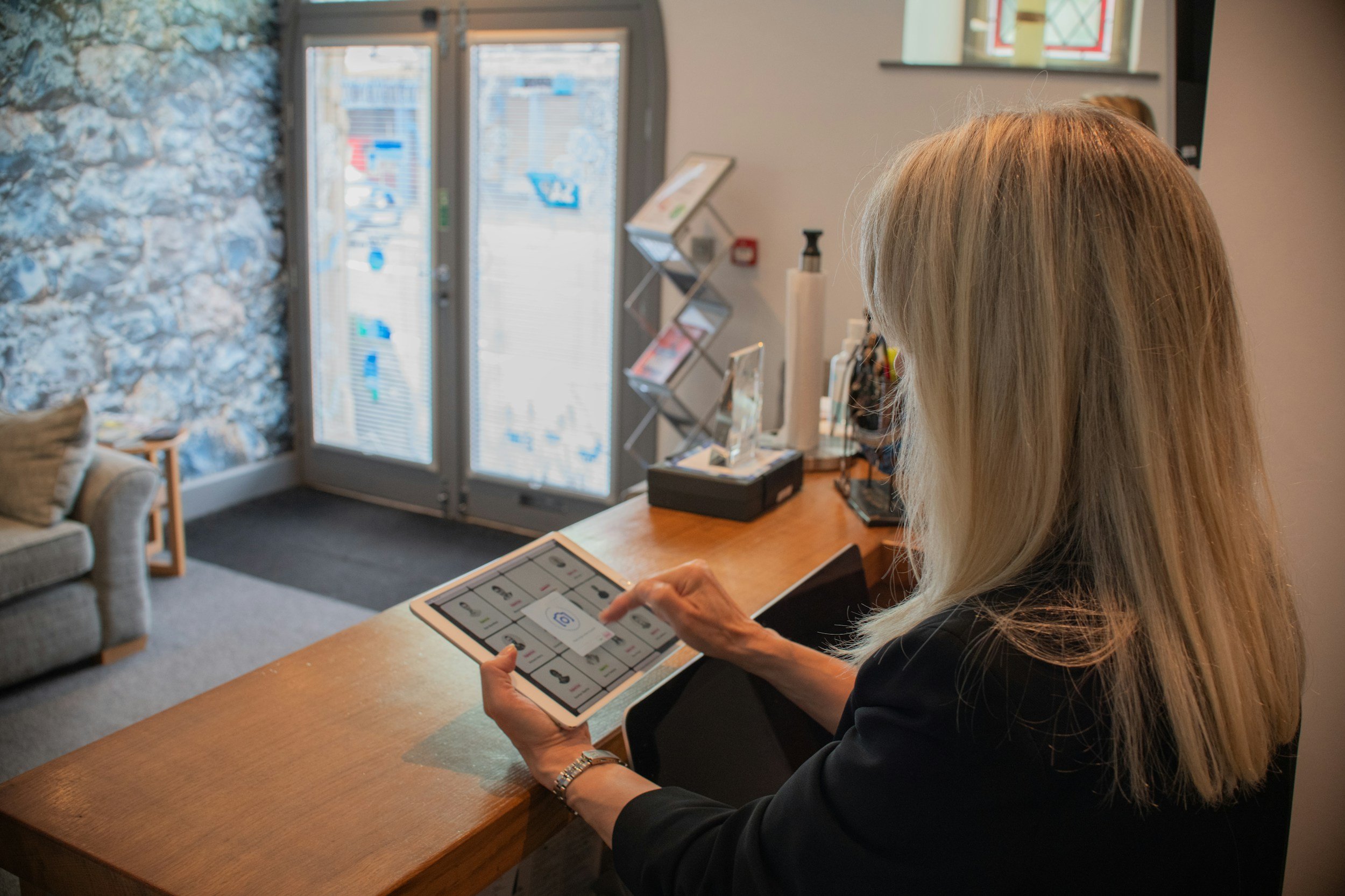 A woman with long blonde hair, wearing a black blazer, sits at a wooden counter inside a modern office or reception area, looking at a tablet device.