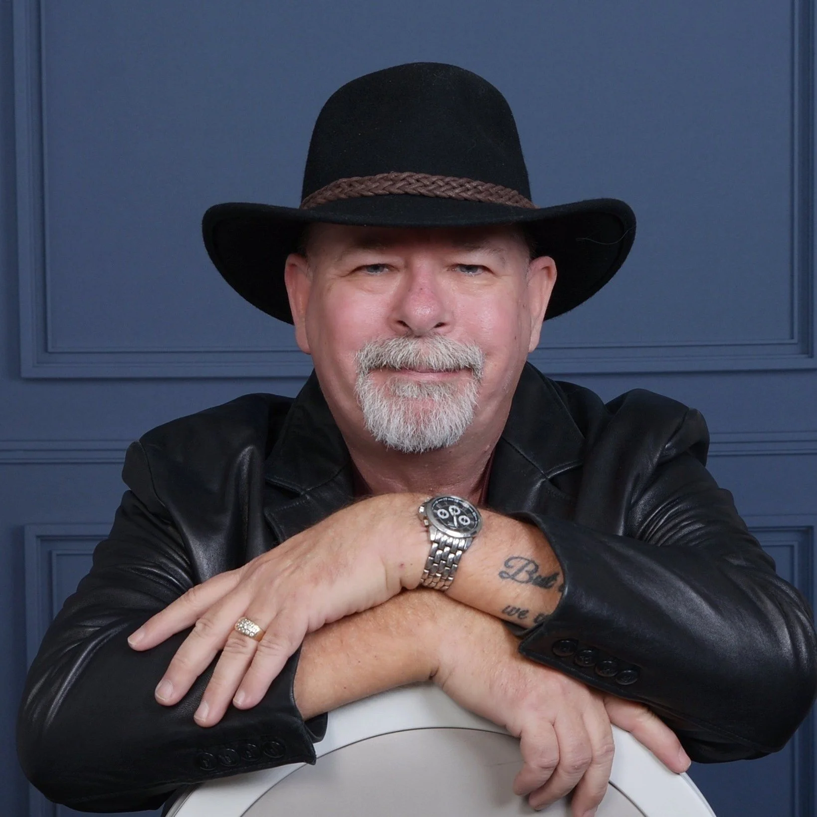 A man with a gray beard wearing a black cowboy hat, black leather jacket, and a silver wristwatch, sitting with arms crossed in front of a blue paneled wall.