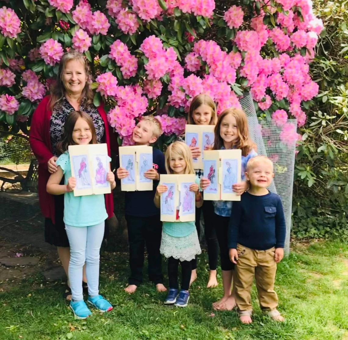 A group of seven children and one woman standing outdoors in front of pink flowering bushes. The children are holding sketchbooks with colored drawings of women in dresses.