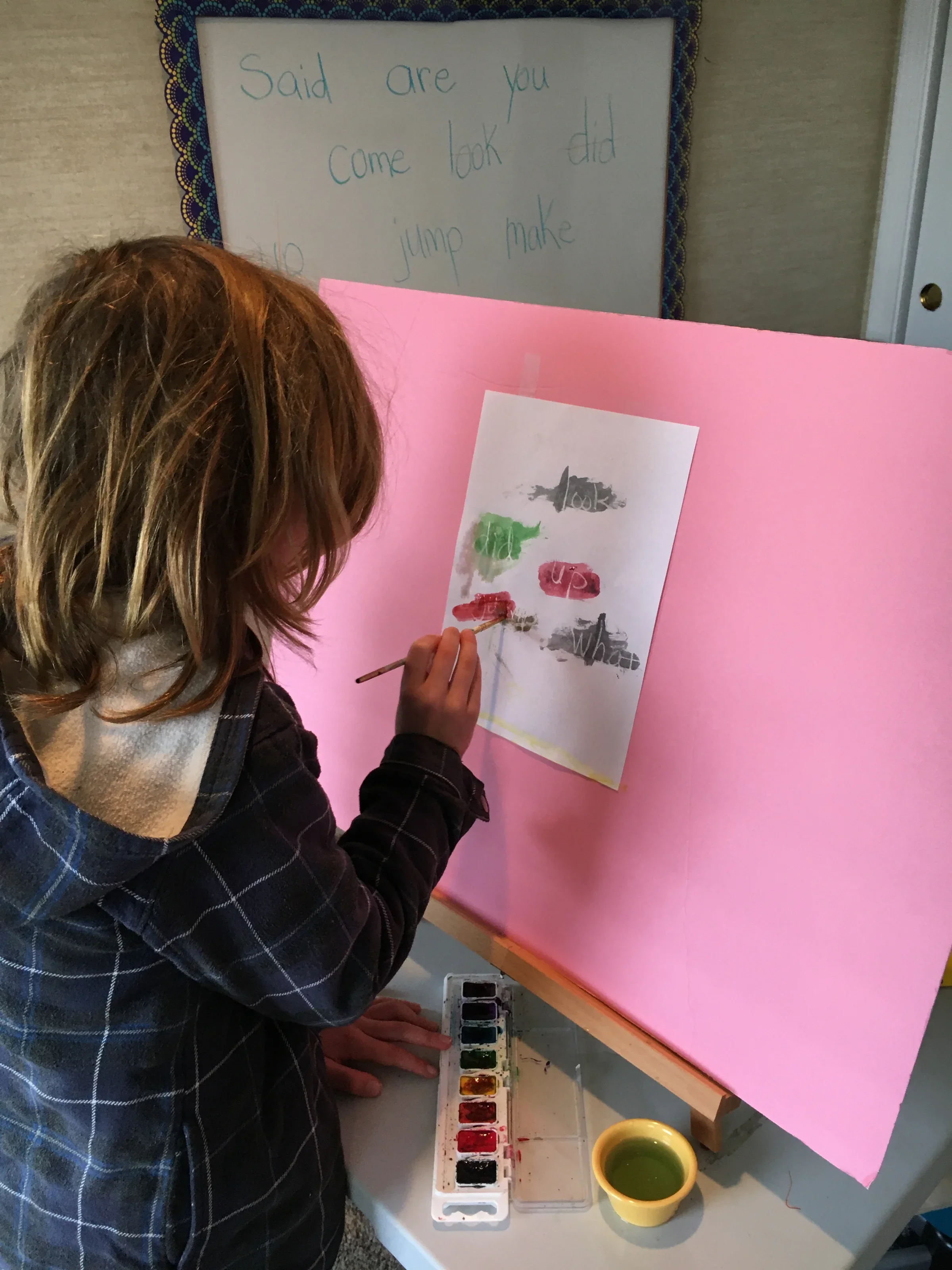 A person with brown hair painting with watercolors at a table. The table has watercolor paints, a cup of water, and pink paper. In the background, a whiteboard with handwritten words is visible.