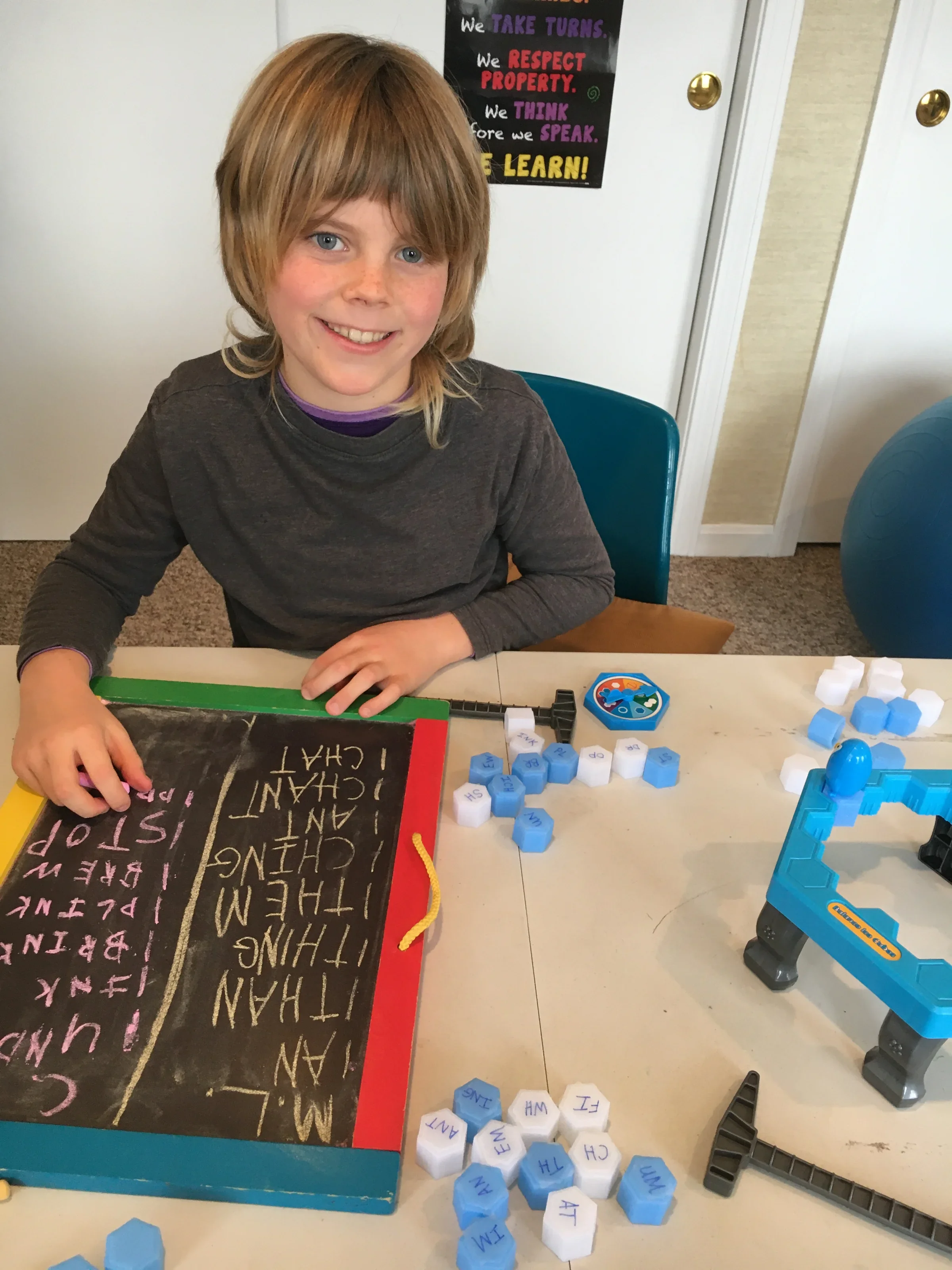 A young boy with blonde hair and blue eyes sitting at a table with educational games and a blackboard. The blackboard has colorful handwritten words about respecting property, kindness, and learning. Spread out on the table are white and blue lettere