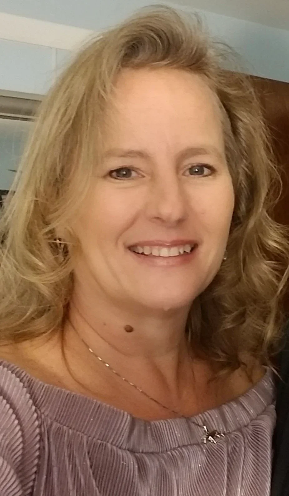 Close-up of a woman with blond, curly hair smiling indoors.