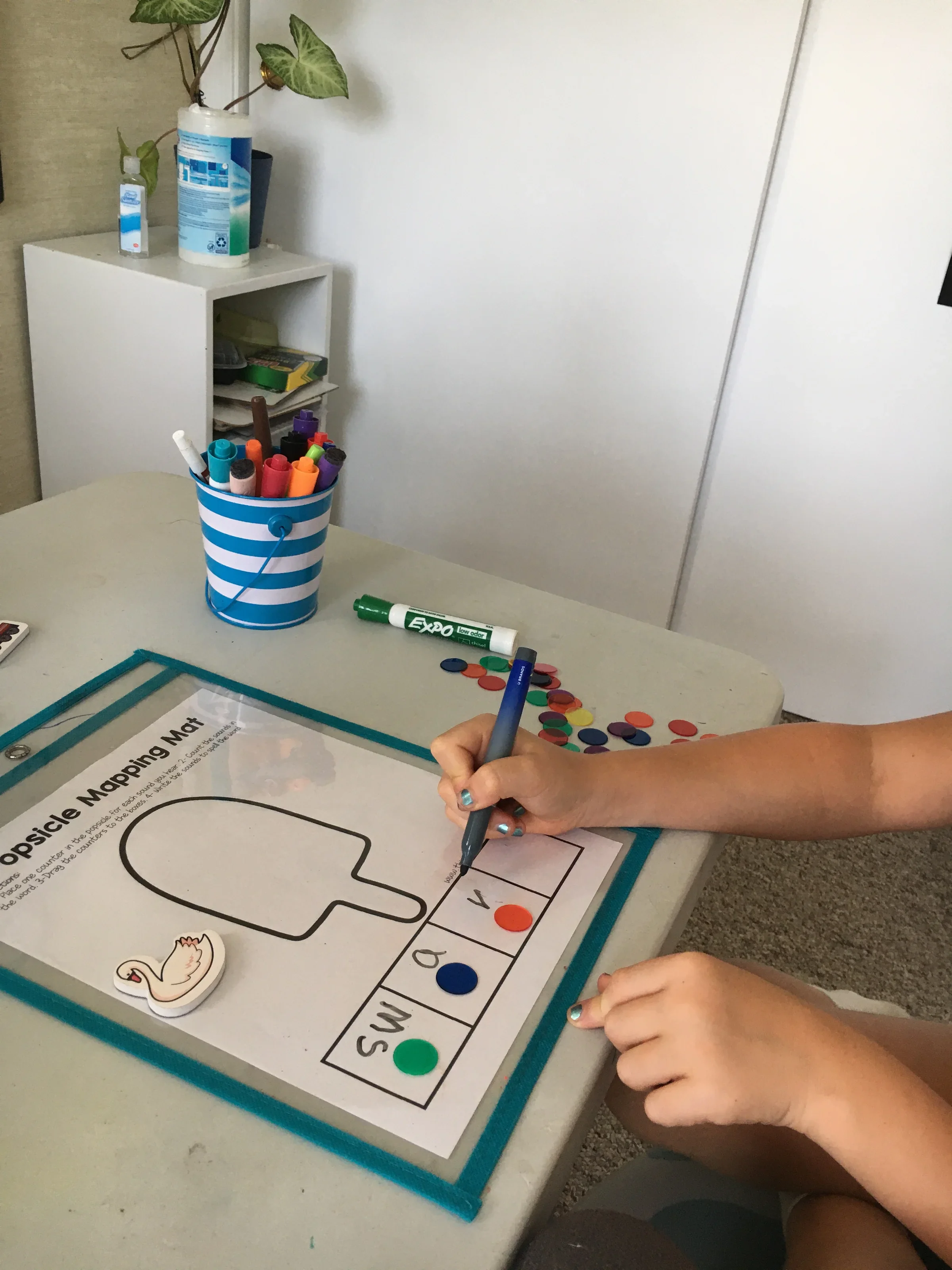 Child coloring a poster about the water cycle with markers, sitting at a table with a supplies tray, a cup of markers, and a small swan sticker, in a room with white walls and shelves.