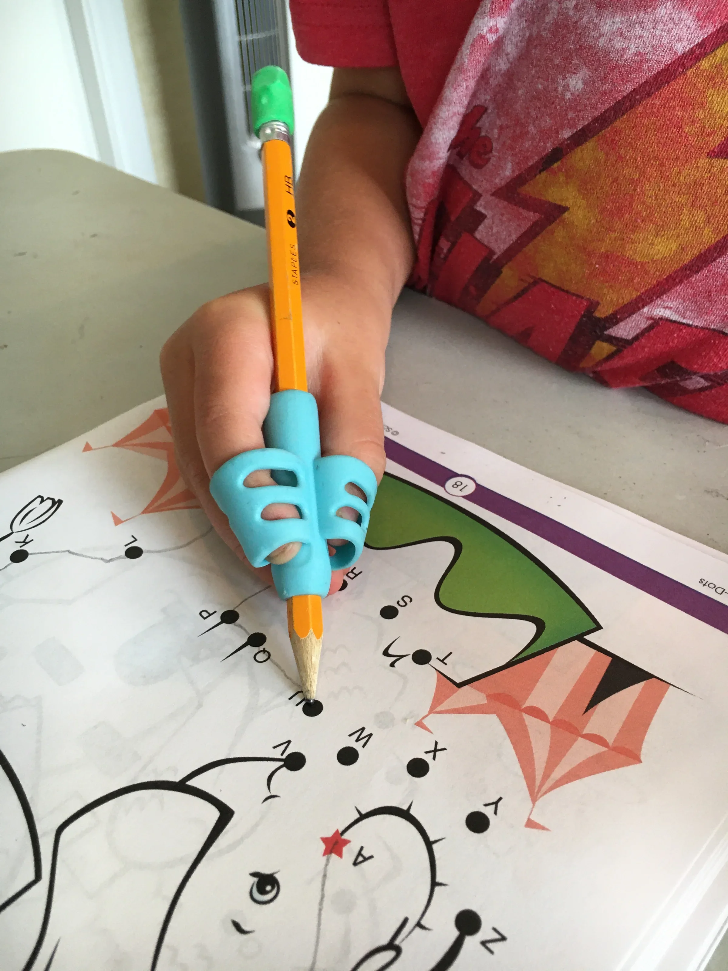 A child's hand holding a yellow pencil with a green eraser, coloring a dot-to-dot activity sheet that features a reindeer with antlers, a red star on its nose, and an orange background with snowflakes and stars.