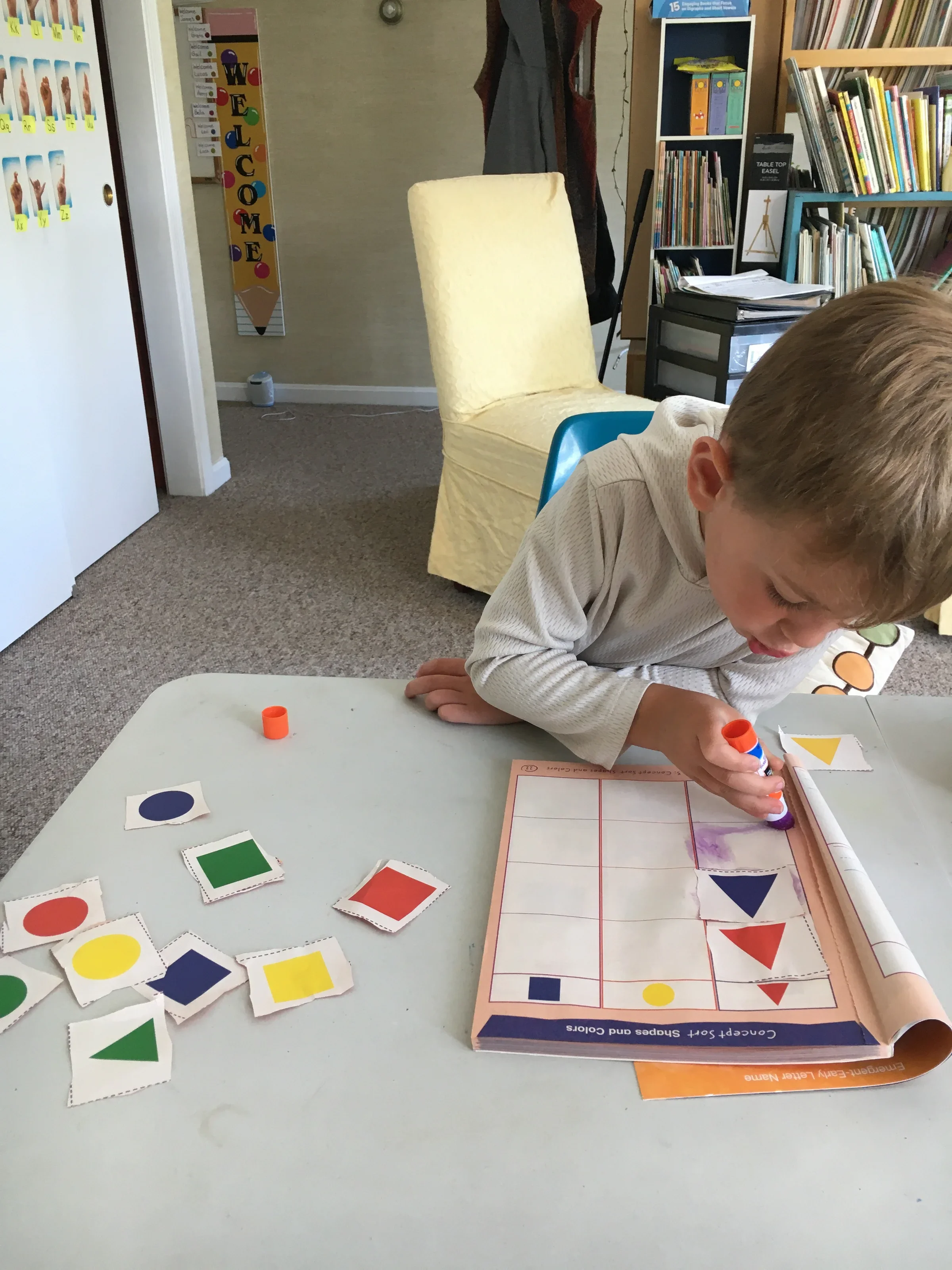 Child with light brown hair working on a colorful shapes matching worksheet at a table, with various colored paper cutouts scattered on the table, in a classroom with bookshelves and educational posters in the background.