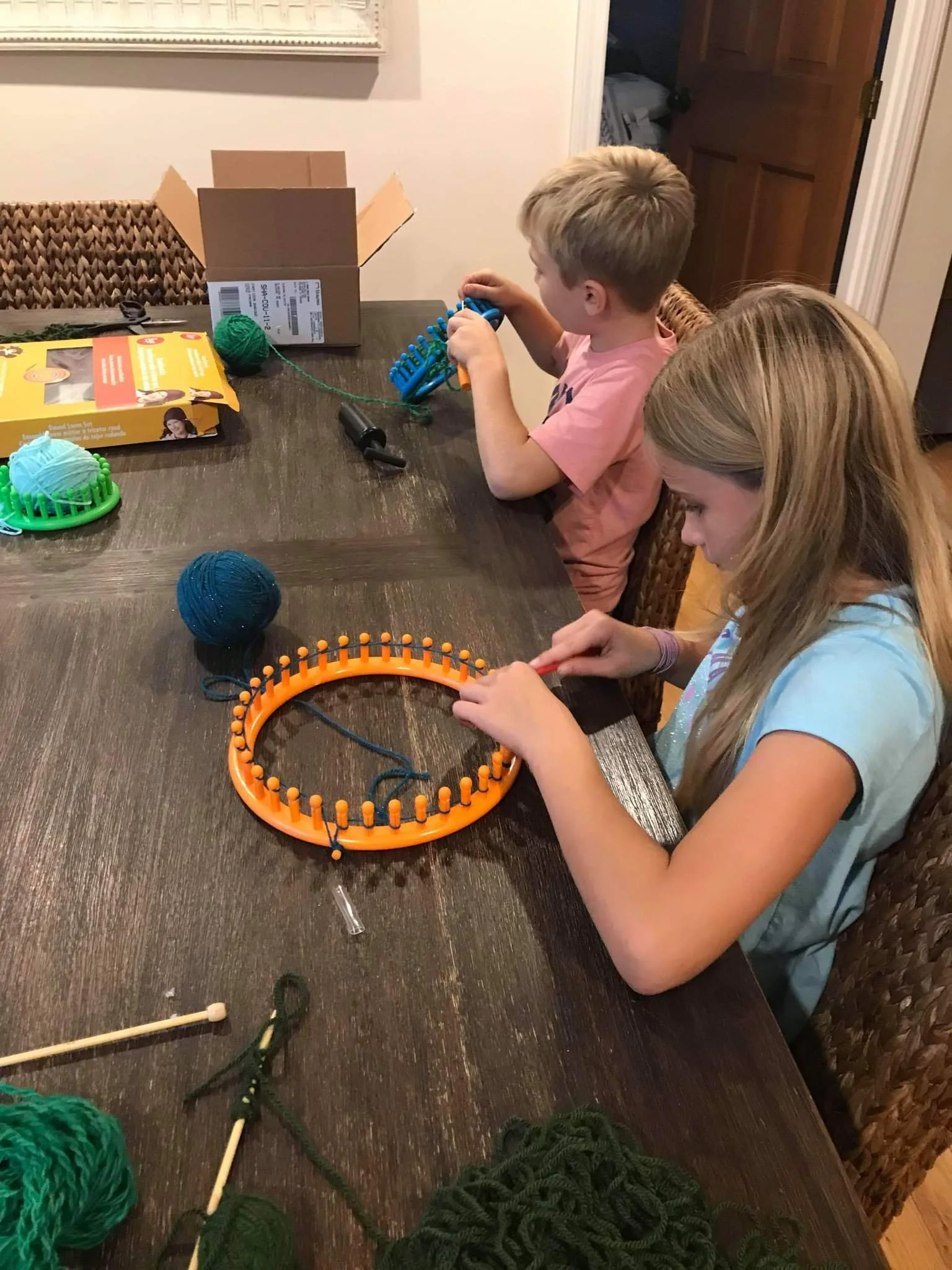 Two children sitting at a wooden table, knitting with yarn and knitting looms. Various balls of yarn and knitting tools are on the table.