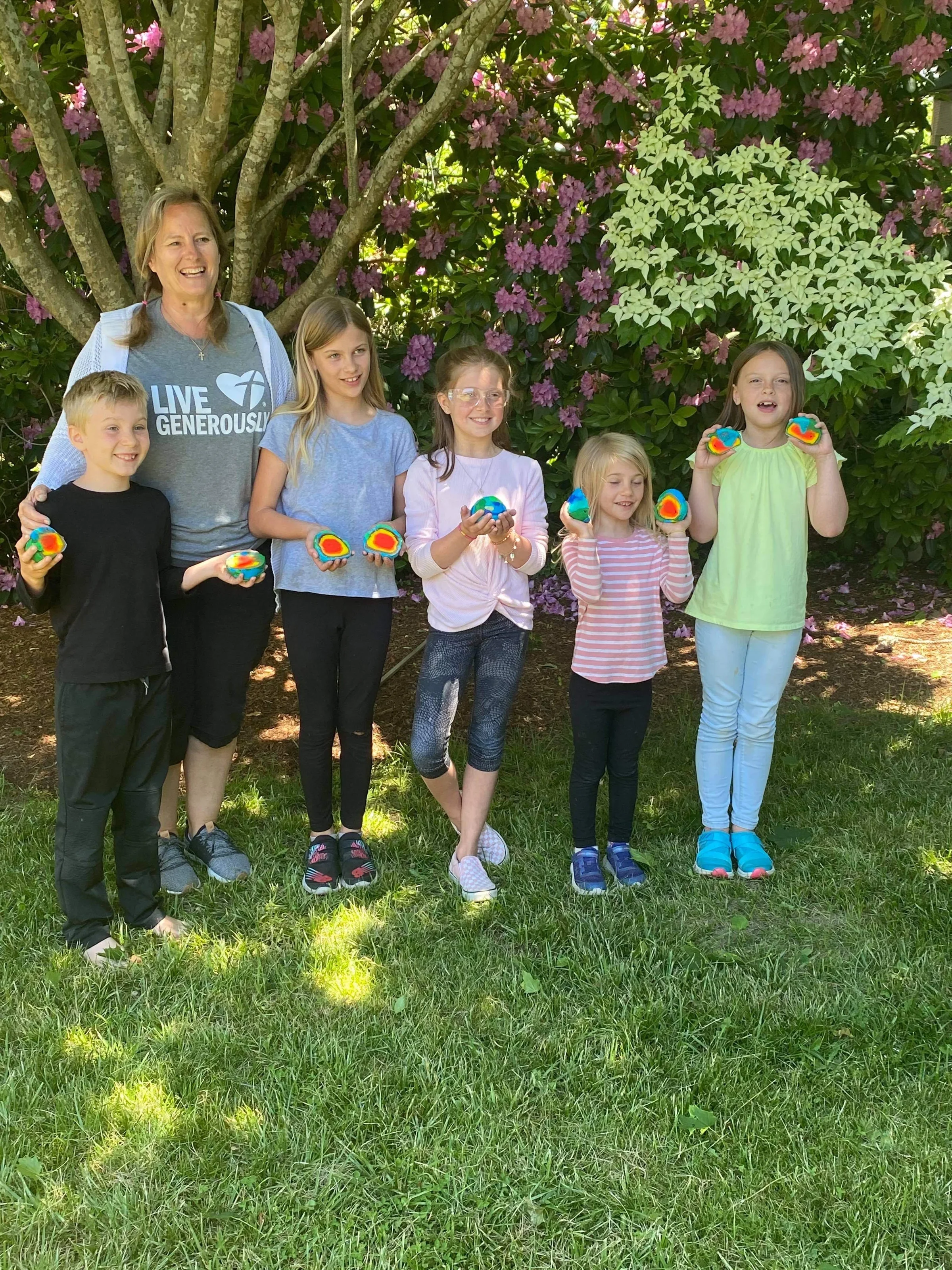 Group of children and a woman standing outdoors in front of a flowering tree, holding colorful rocks and smiling.