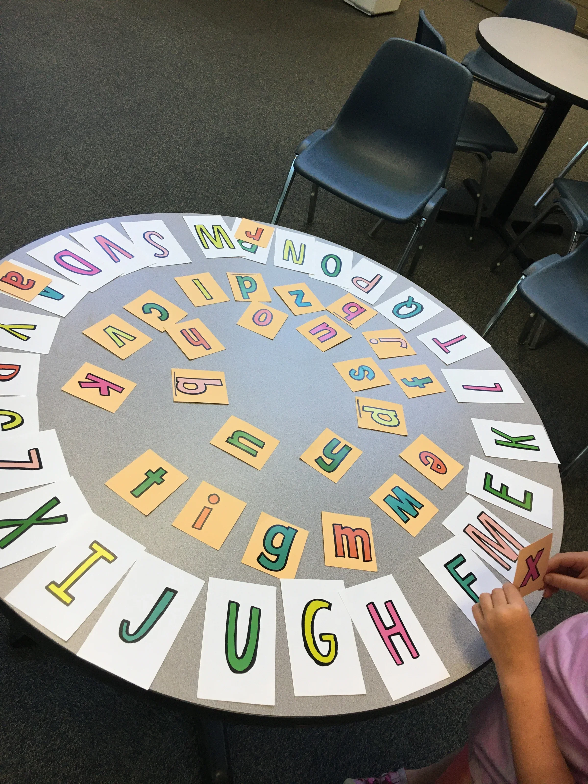 Colorful alphabet flashcards arranged in a semi-circle on a table with a child's hand reaching for a card.