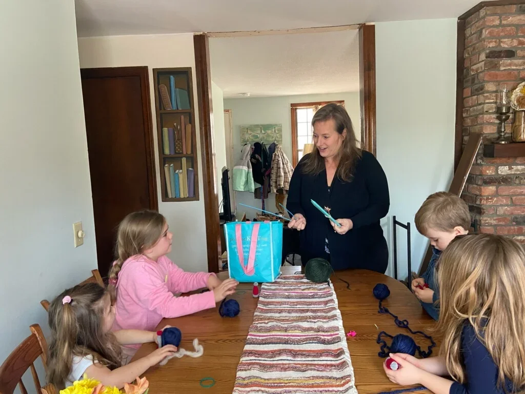 A woman standing at a dining table with four children, guiding them in a craft activity with yarn and scissors, in a cozy home interior.