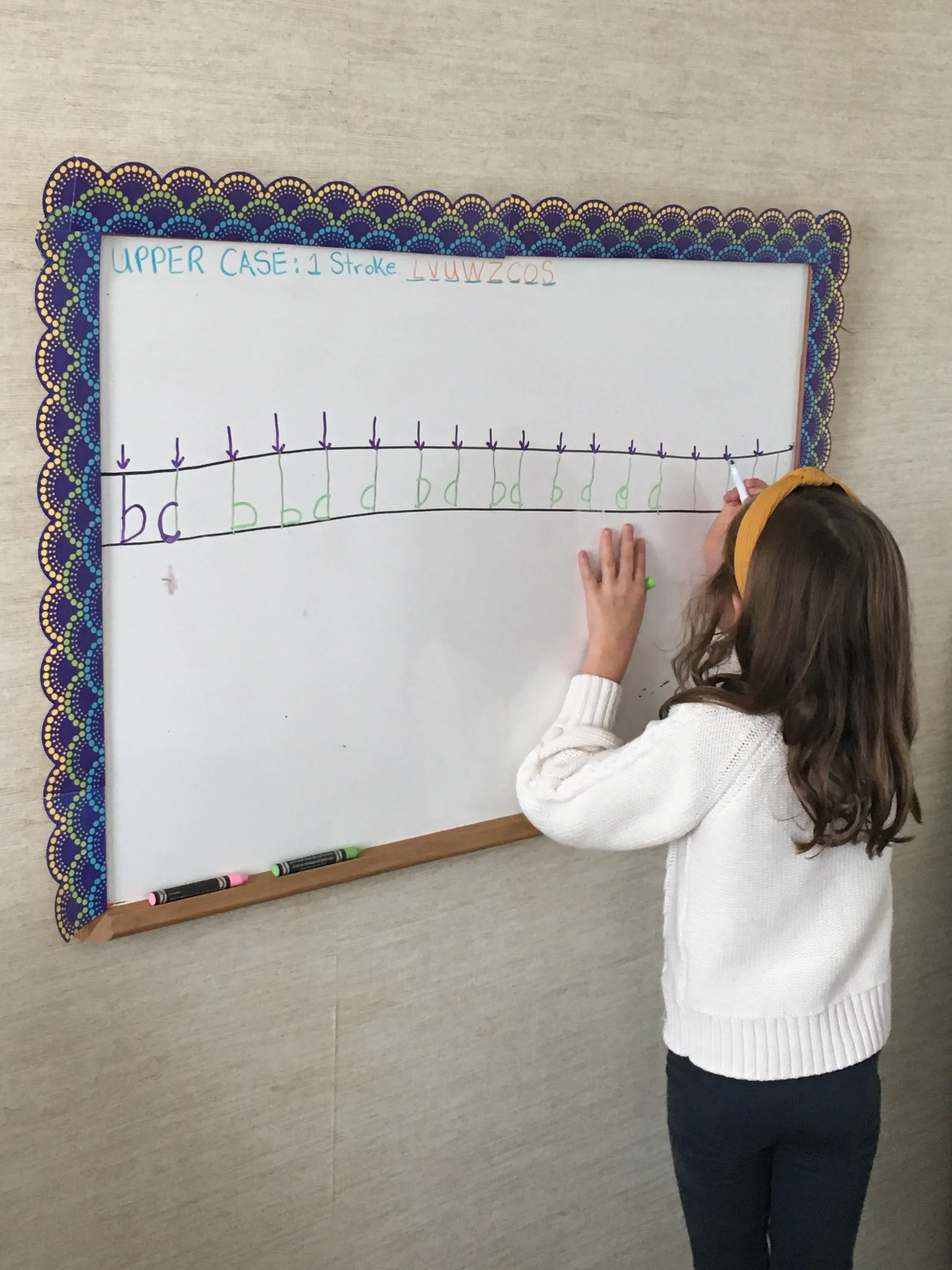 A young girl with long, wavy hair writing on a whiteboard with markers. The whiteboard has a decorative border and some handwritten notes at the top, including 'UPPER CASE: 1 stroke' and a sequence of letters. The girl is wearing a white sweater and 