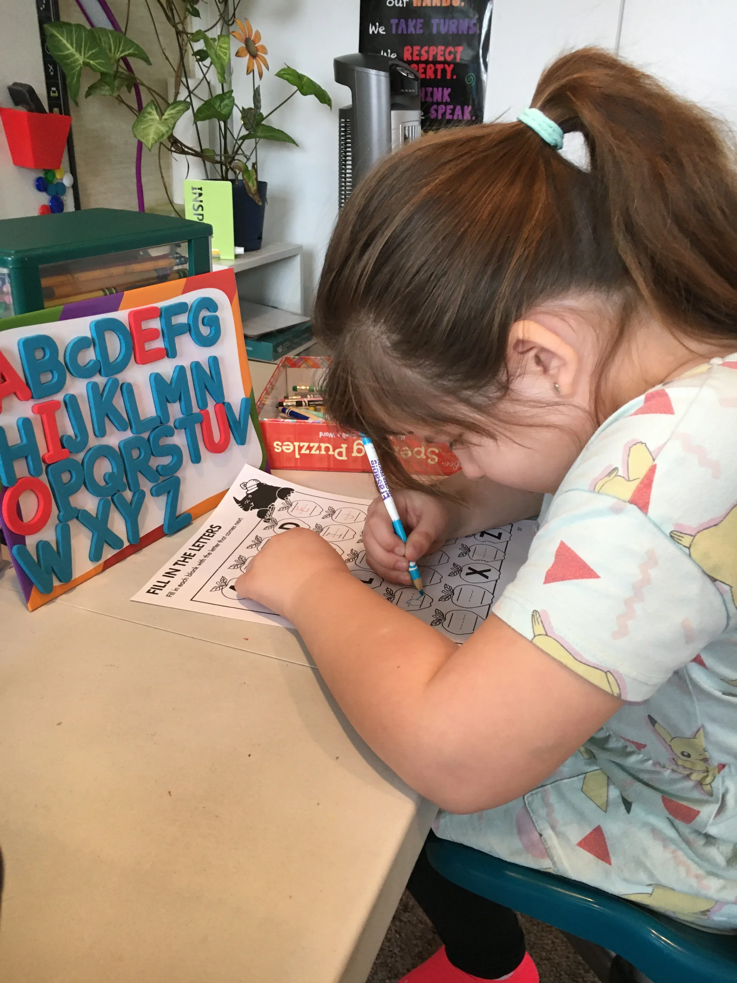 A young girl with striped basketball shorts, a colorful unicorn shirt, and red socks is writing on a worksheet at a desk. In front of her is an educational foam alphabet board with colorful letters, a box of crayons, and a book. Behind her, there is 