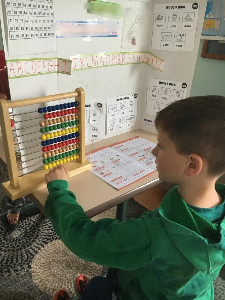 A boy wearing a green hoodie playing with an abacus on a classroom desk, with educational posters and alphabet strip on the wall behind him.