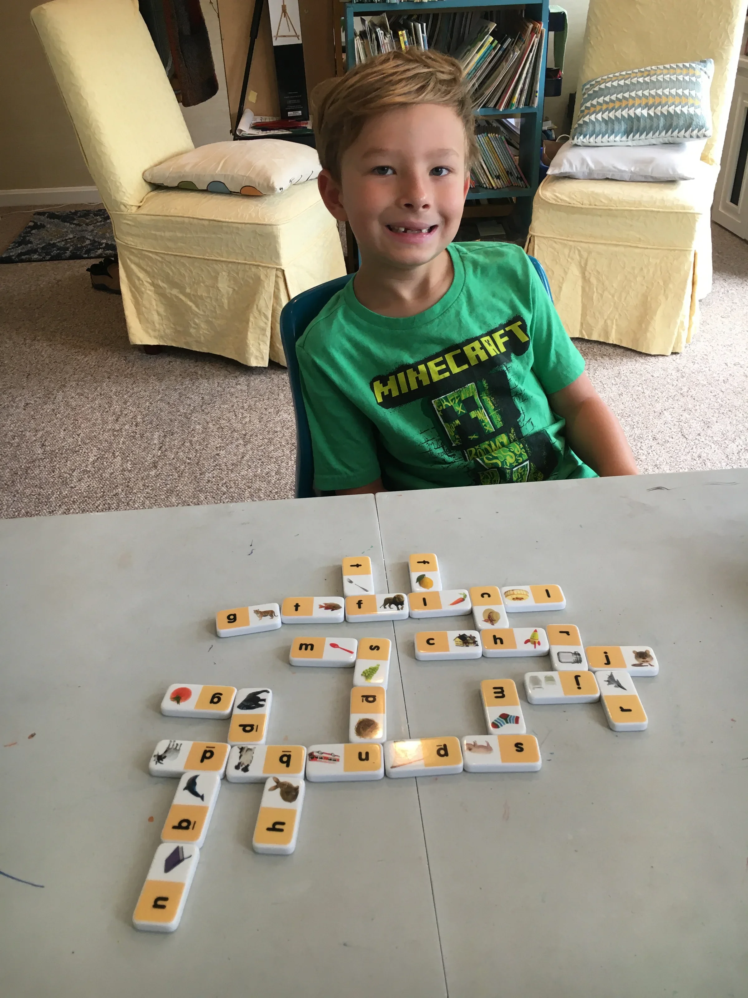 A young boy with short, light brown hair is sitting at a table with a set of educational tiles, which have images and letters on them, laid out in front of him. Behind him, there are two yellow armchairs with pillows, a bookshelf filled with books, a